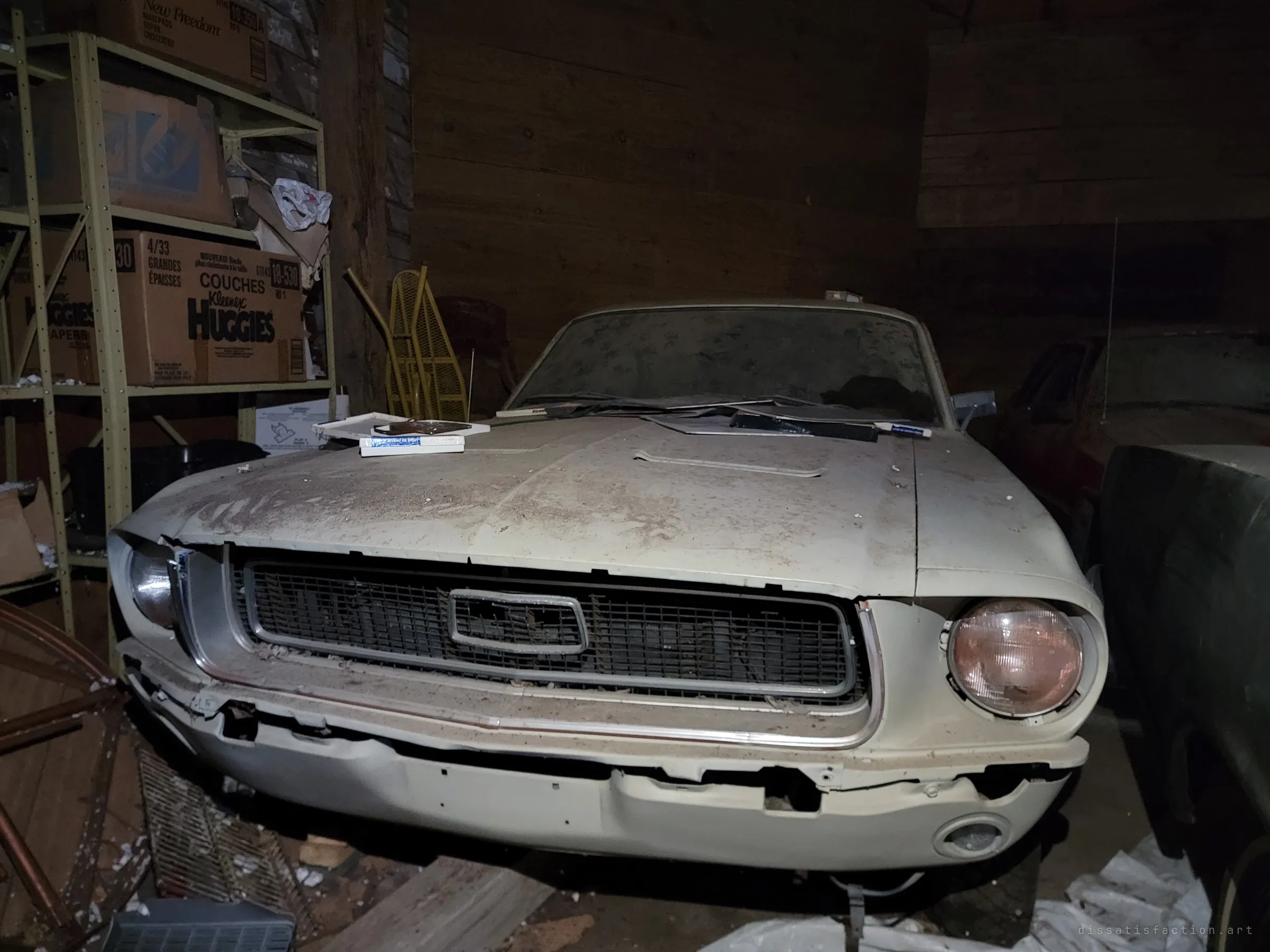 A dust-caked cream 1960s muscle car sits nose-first in a dark wooden barn, papers and boxes left on its hood beside shelves of old cardboard boxes. The layer of grime shows how long it has been parked and forgotten.