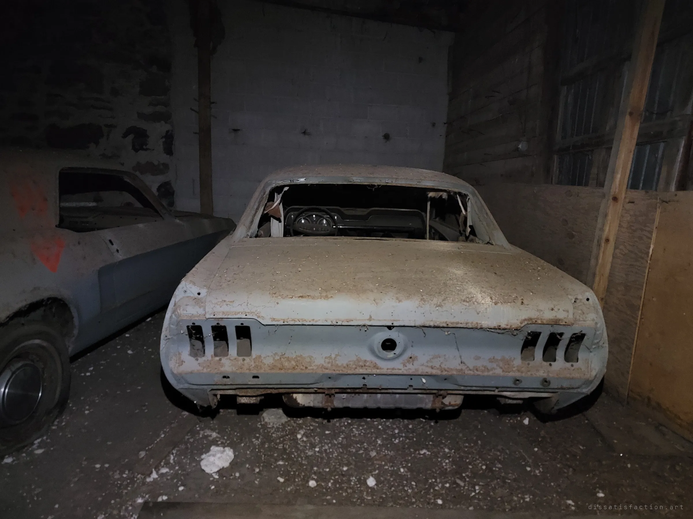 The rusted rear of a stripped classic coupe with its back window gone and tail-light housings empty, parked against a cinder-block wall beside another primer-grey car. Rust blooms across the trunk lid and bumper line.