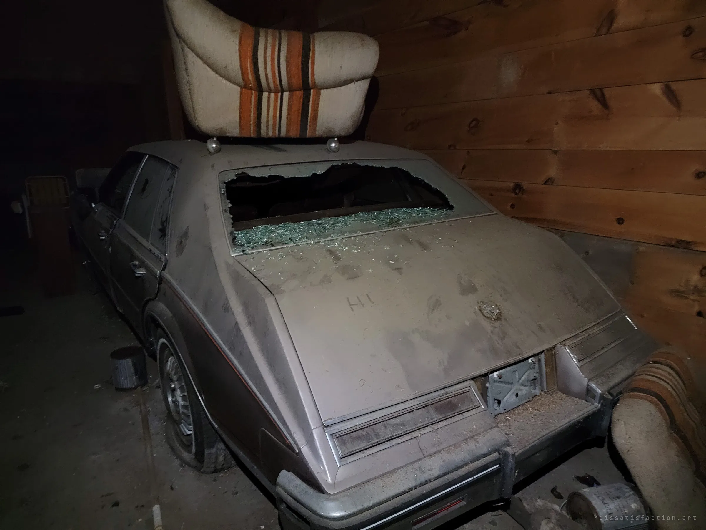 A dusty grey luxury sedan with a smashed rear window and a striped car seat balanced on its roof, wedged against pine-panelled walls. Someone has fingered a greeting into the dust on the trunk.
