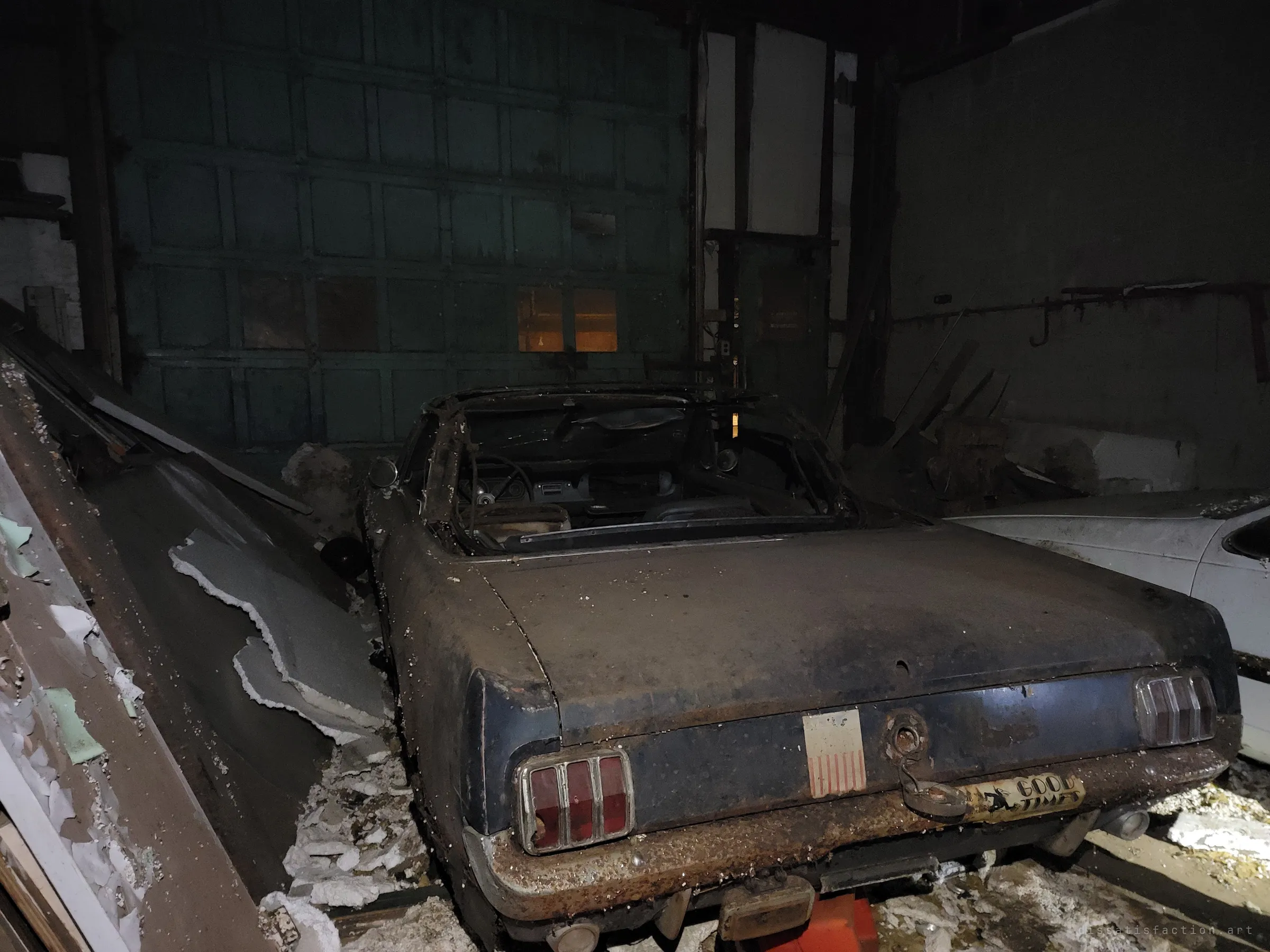 A rust-pitted dark convertible on jack stands in front of a large sectional garage door, collapsed panels and debris piled along its side. A faded flag decal and a novelty plate still cling to the rear.