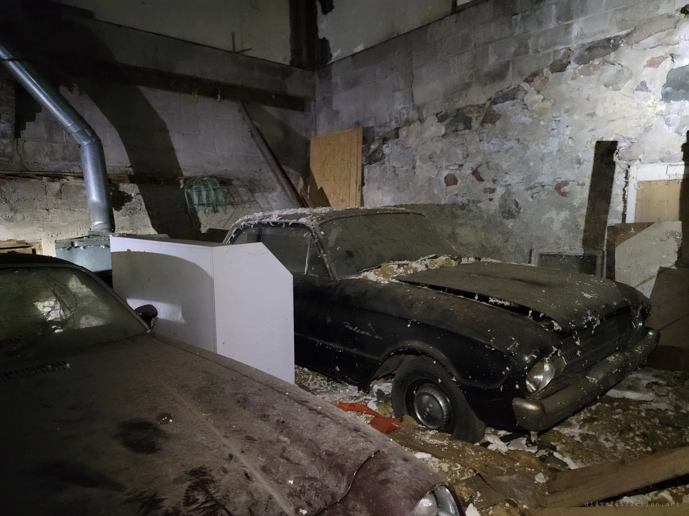A black early-1960s compact sedan sits on a flat tire against a crumbling stone and block wall, insulation scattered over its hood. The mix of old fieldstone and patched masonry frames the car like a relic.