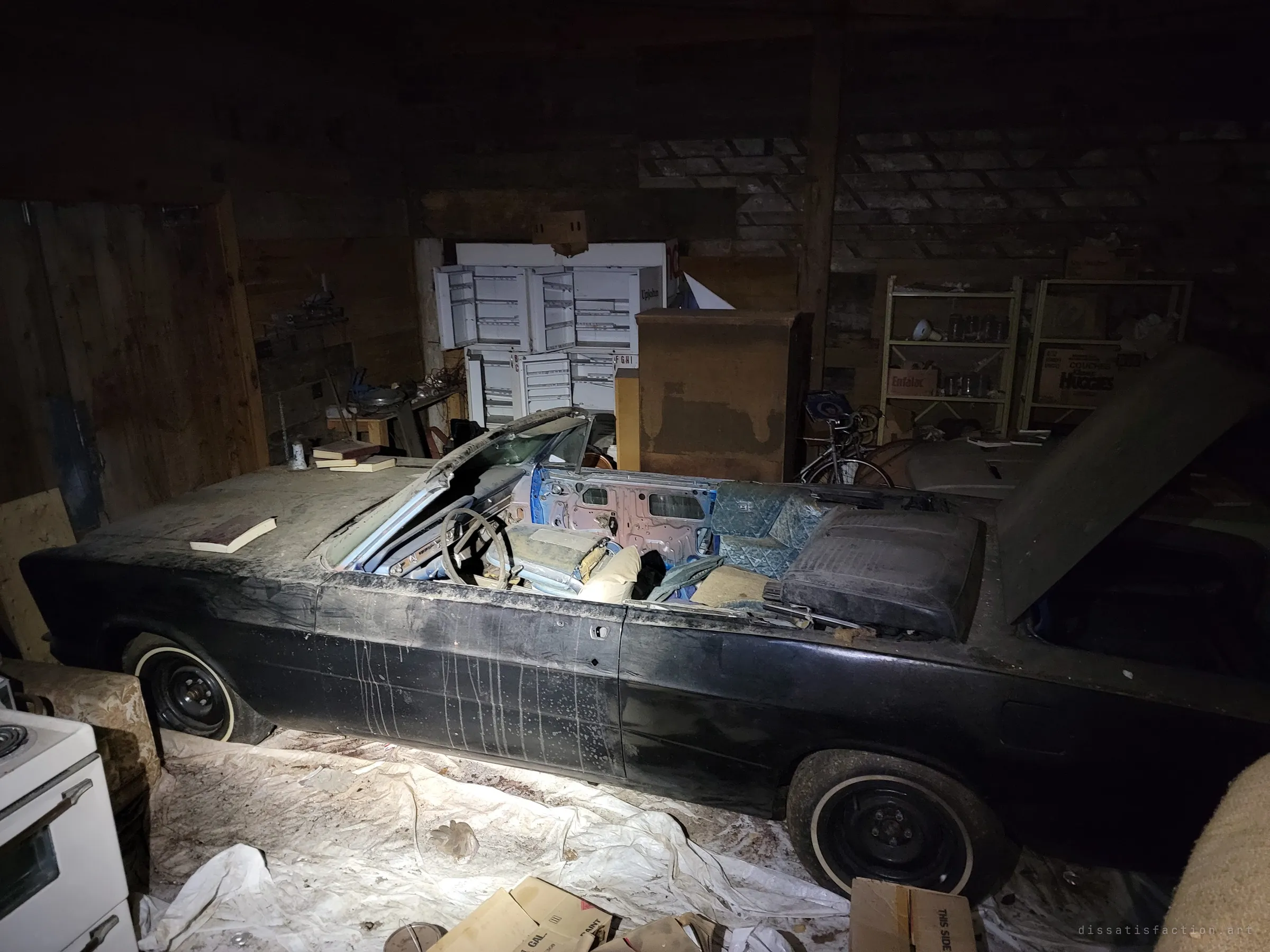A black convertible with its top gone and seats torn apart, trunk lid raised, parked on plastic sheeting in a cluttered barn full of cabinets, shelves and a bicycle. Old books lie scattered on its rear deck.
