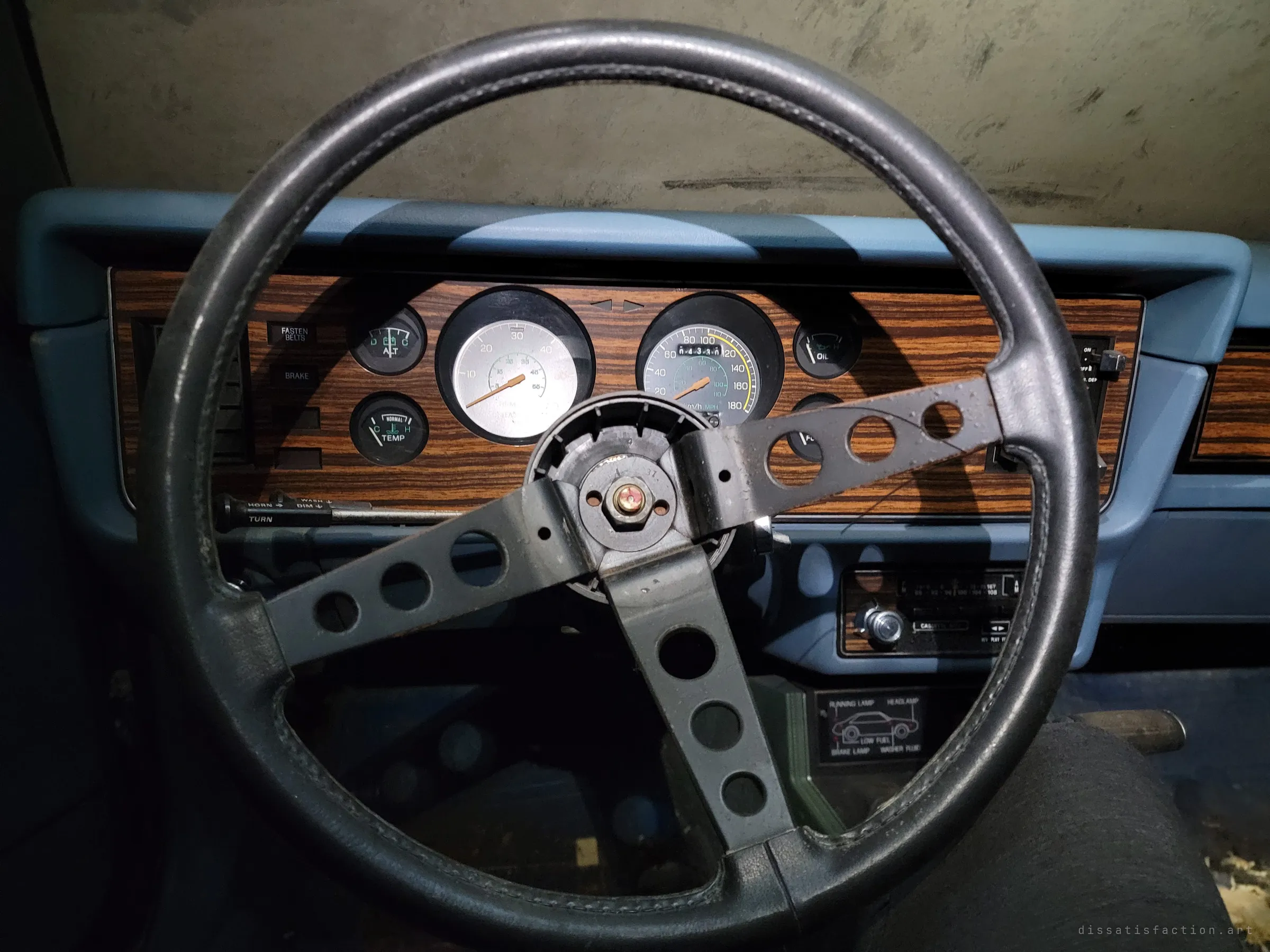Driver's view through a drilled three-spoke aftermarket steering wheel onto a woodgrain gauge cluster and pale blue dash. The period cassette deck and warning-light panel date the car to the early eighties.