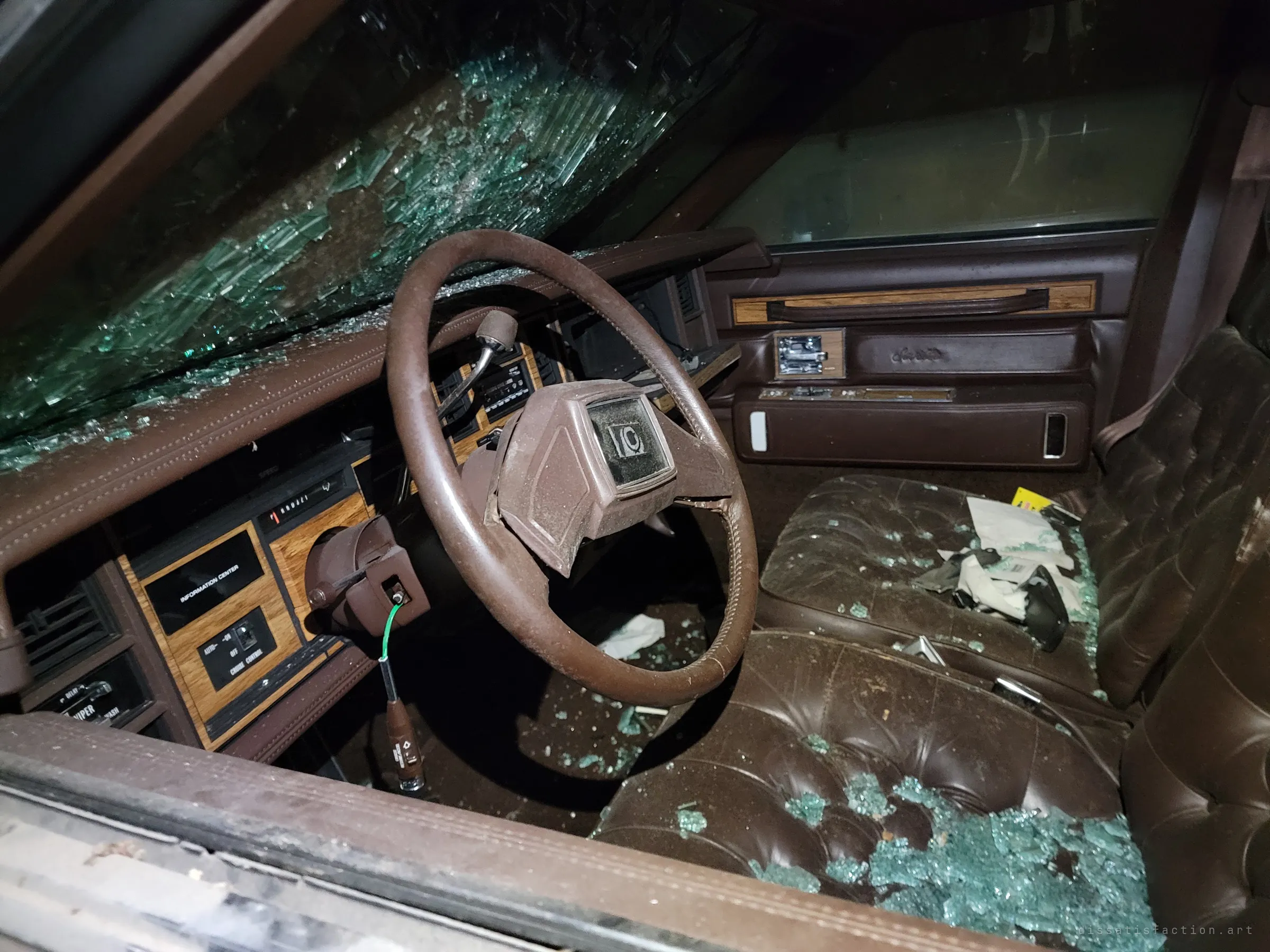 A brown luxury coupe's cabin, its windshield caved in and tufted leather seats covered in shattered safety glass. Loose papers and debris lie on the passenger seat, showing how far the car has been left to decay.