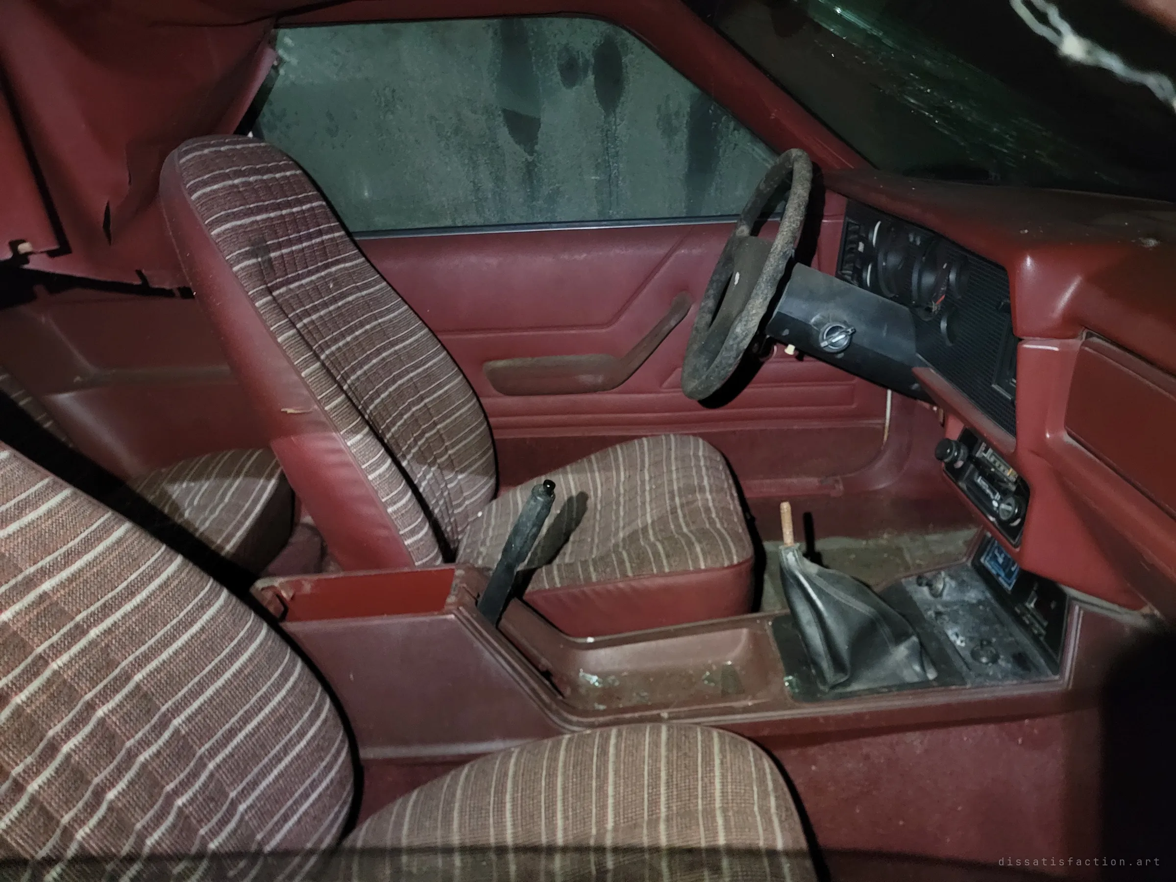 The burgundy interior of an eighties hatchback, with striped cloth bucket seats, a manual shifter and a grimy steering wheel. Mould streaks the side window, a sign of long storage in a damp building.