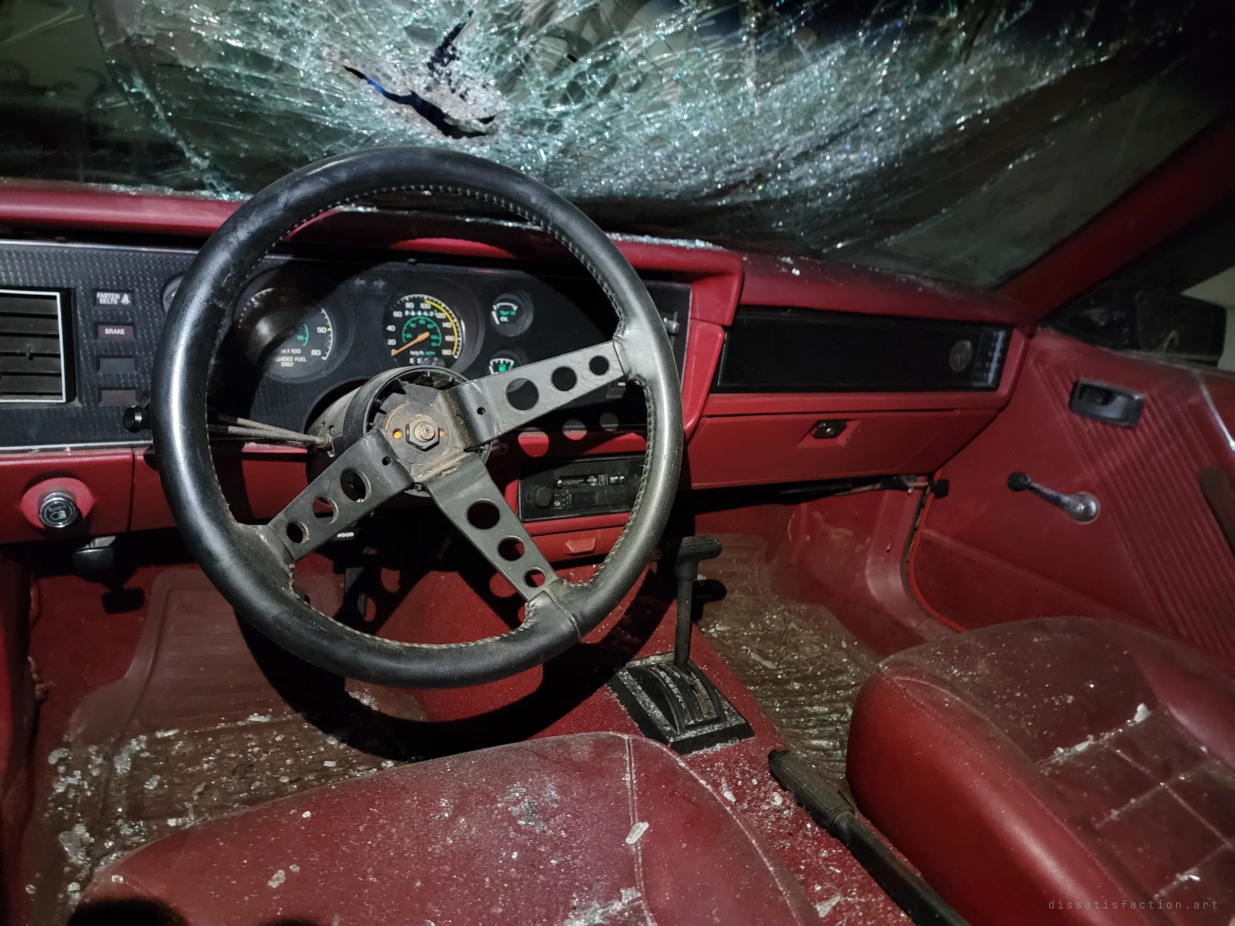 A red interior behind a drilled black steering wheel, with the windshield smashed through in a spiderweb of cracks. Glass fragments are scattered over the seats and carpet.