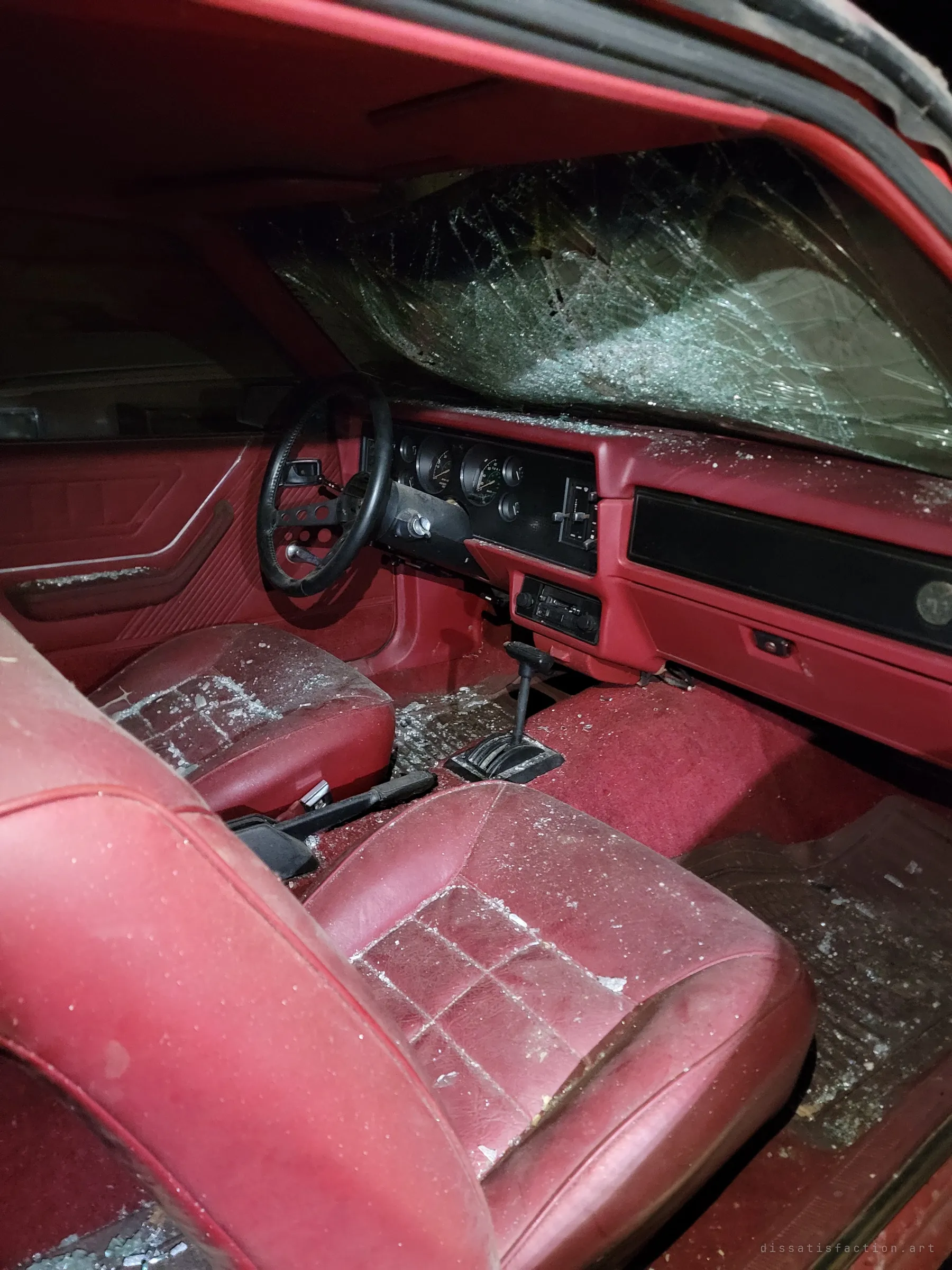 The same red car seen from the passenger door, its windshield shattered and red vinyl seats dusted with glass. The black dash and automatic shifter sit under the broken glass.