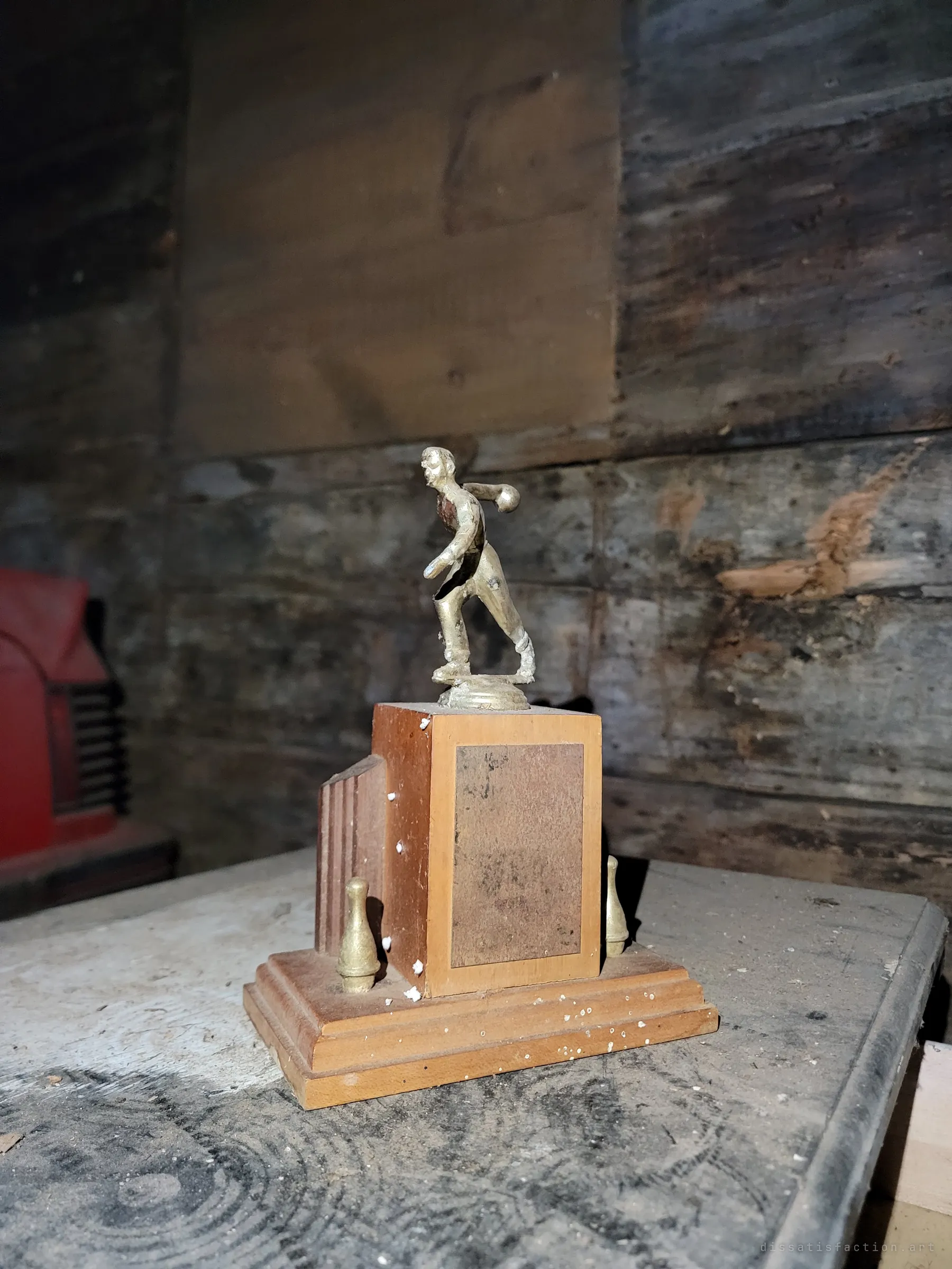 A small bowling trophy with a brass bowler figure and two pins, its engraved plate worn blank, left on a dusty bench against weathered barn boards. A red car's grille peeks in from the edge, a trace of someone's life among the stored cars.