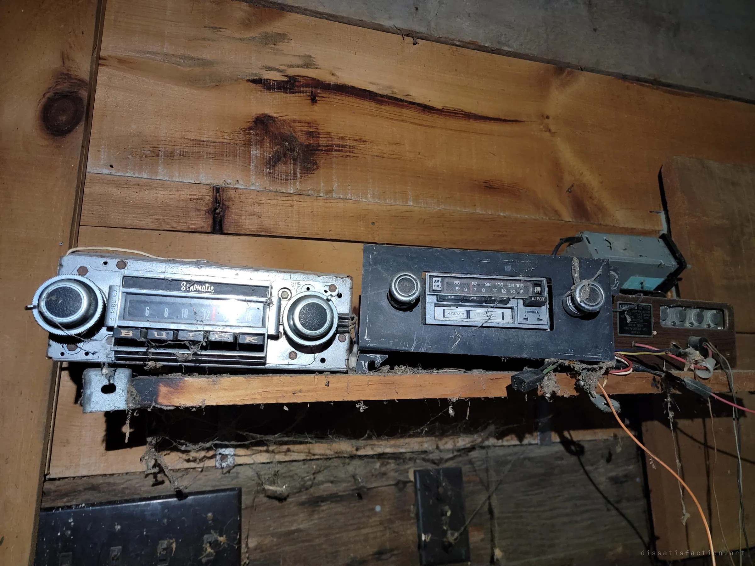 A row of salvaged car radios on a cobwebbed wooden shelf, from a chrome push-button unit to an eighties cassette deck, with loose wiring hanging below. A small archive of dashboard audio kept by the collector.