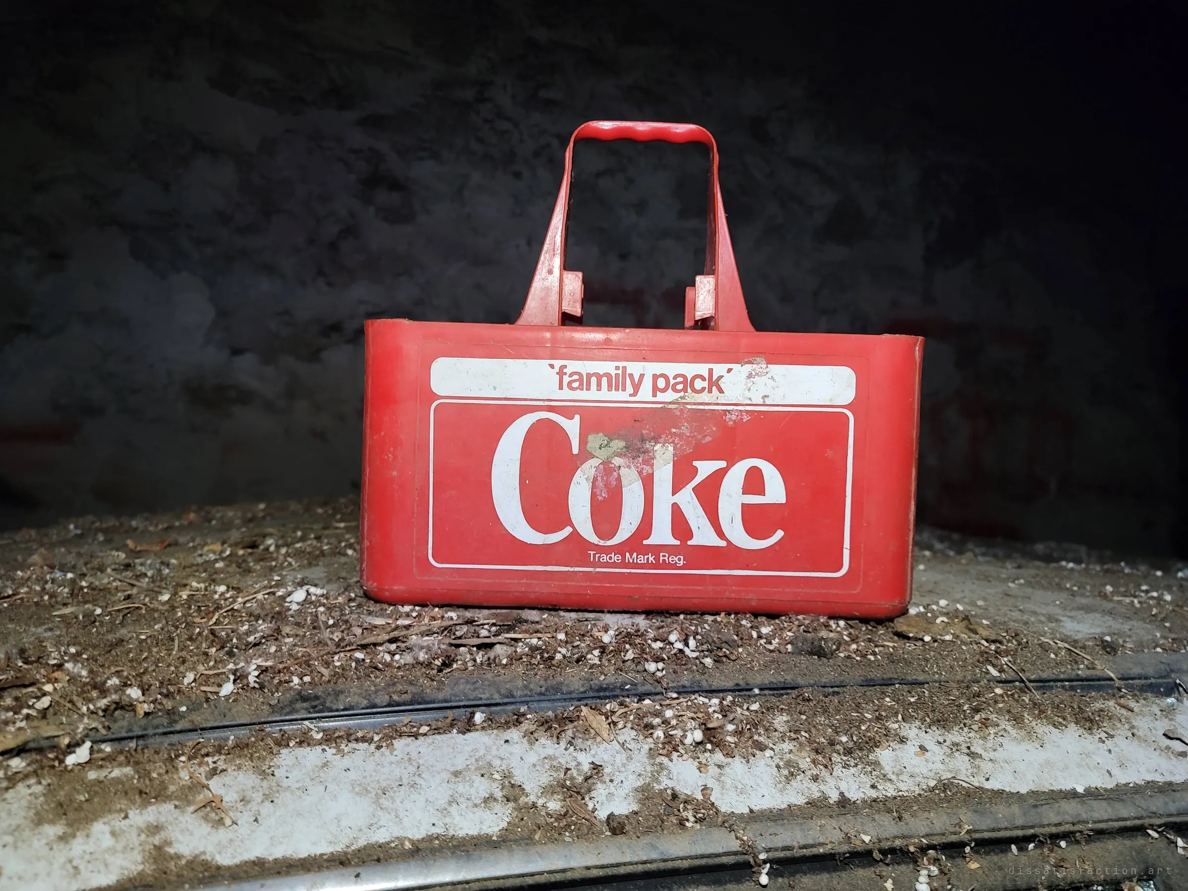 A red plastic soft-drink carrier with period branding sits on a dirt-covered car roof or hood in the dark. Debris and droppings around it show how long nothing has moved here.