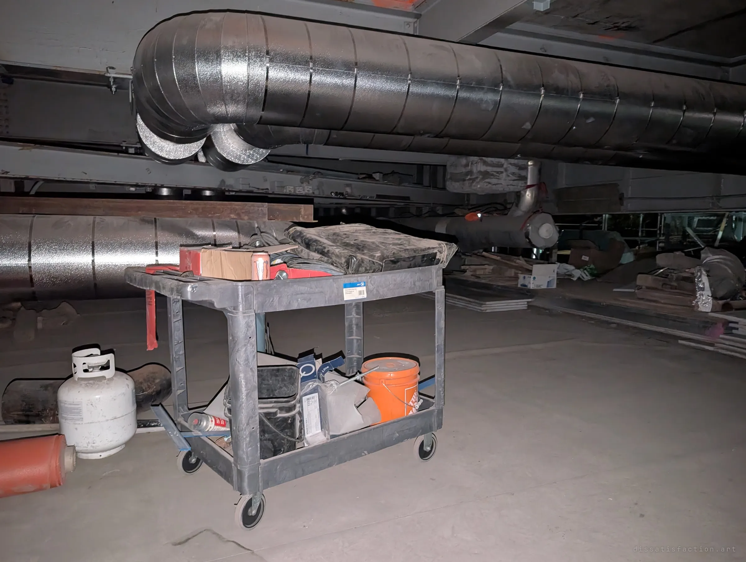 A utility cart loaded with tools, a bucket and a propane tank sits in a low, dusty void beneath large foil-wrapped ducts. Workers' gear left mid-job in the crawlspace under the structure.