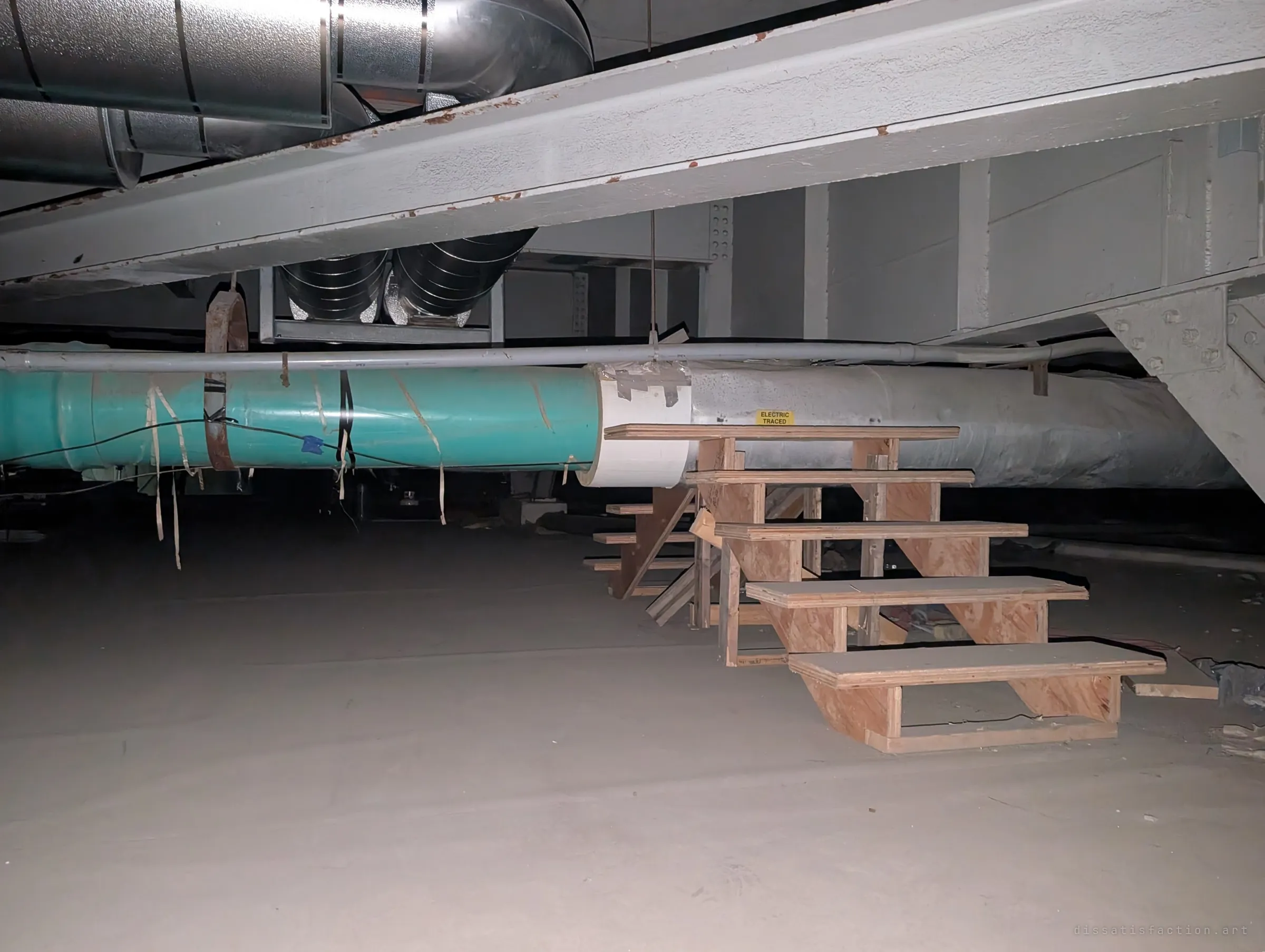 A makeshift plywood staircase climbs over a teal-wrapped pipe beneath painted steel girders and silver ductwork. Improvised stairs are the only way across the services in the crawlspace.