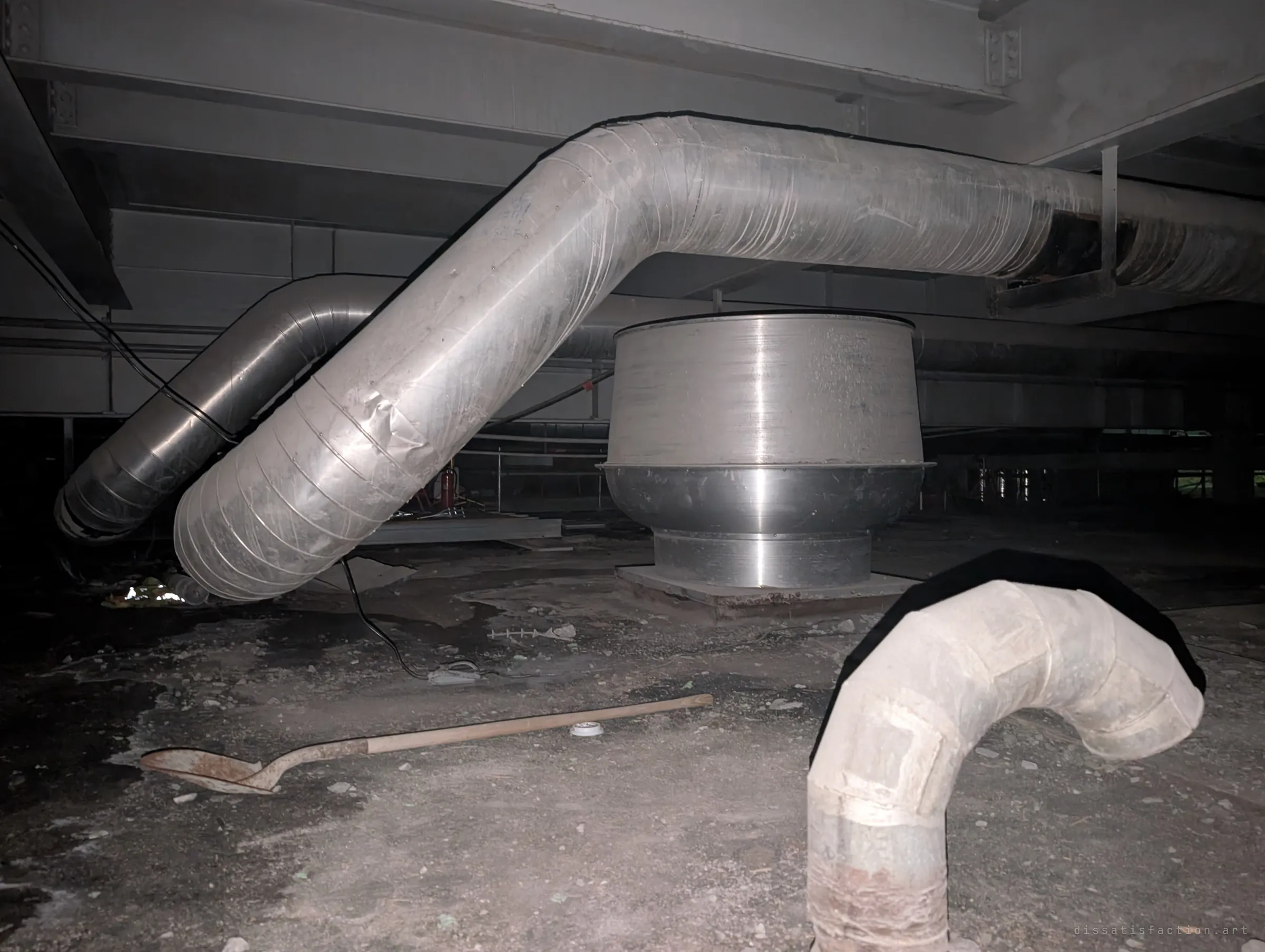 Bent insulated ducts snake around a large round metal housing on a debris-strewn floor, with an old shovel lying in the dust. The service level feels long abandoned despite the working equipment.