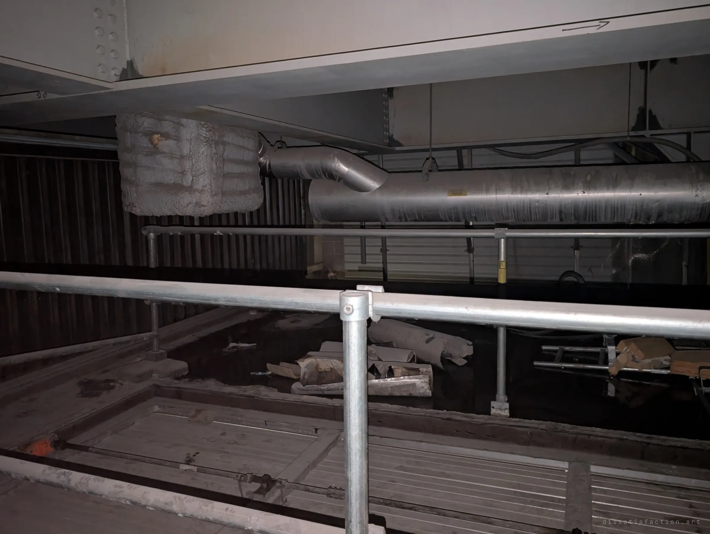 Galvanised scaffold railings guard an opening in the floor, with torn insulation and debris in the pit below and ducts running under the steel above. Part of the underside is opened up for works.