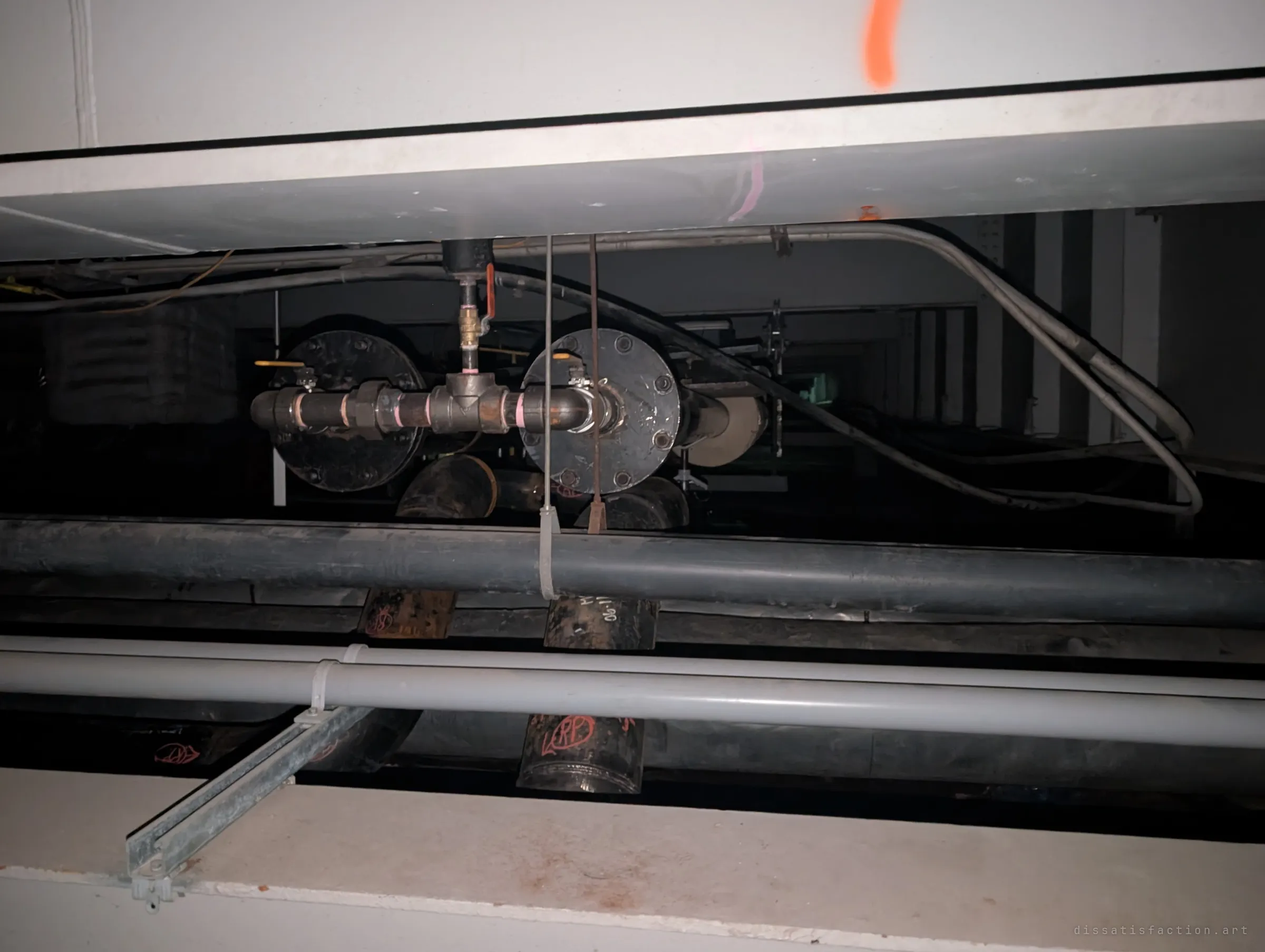 Through a slot between two white girders, flanged pipe fittings and valves sit among dark conduits and hangers. A close look at the plumbing packed into the structure.