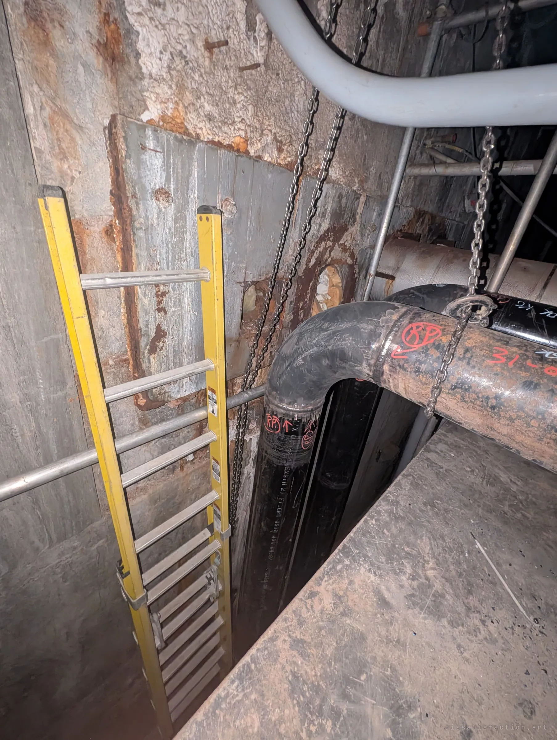 A yellow fibreglass ladder drops into a rusted concrete shaft beside new black pipes marked with red inspection stamps and hung on chains. It shows the way down to the lower level.