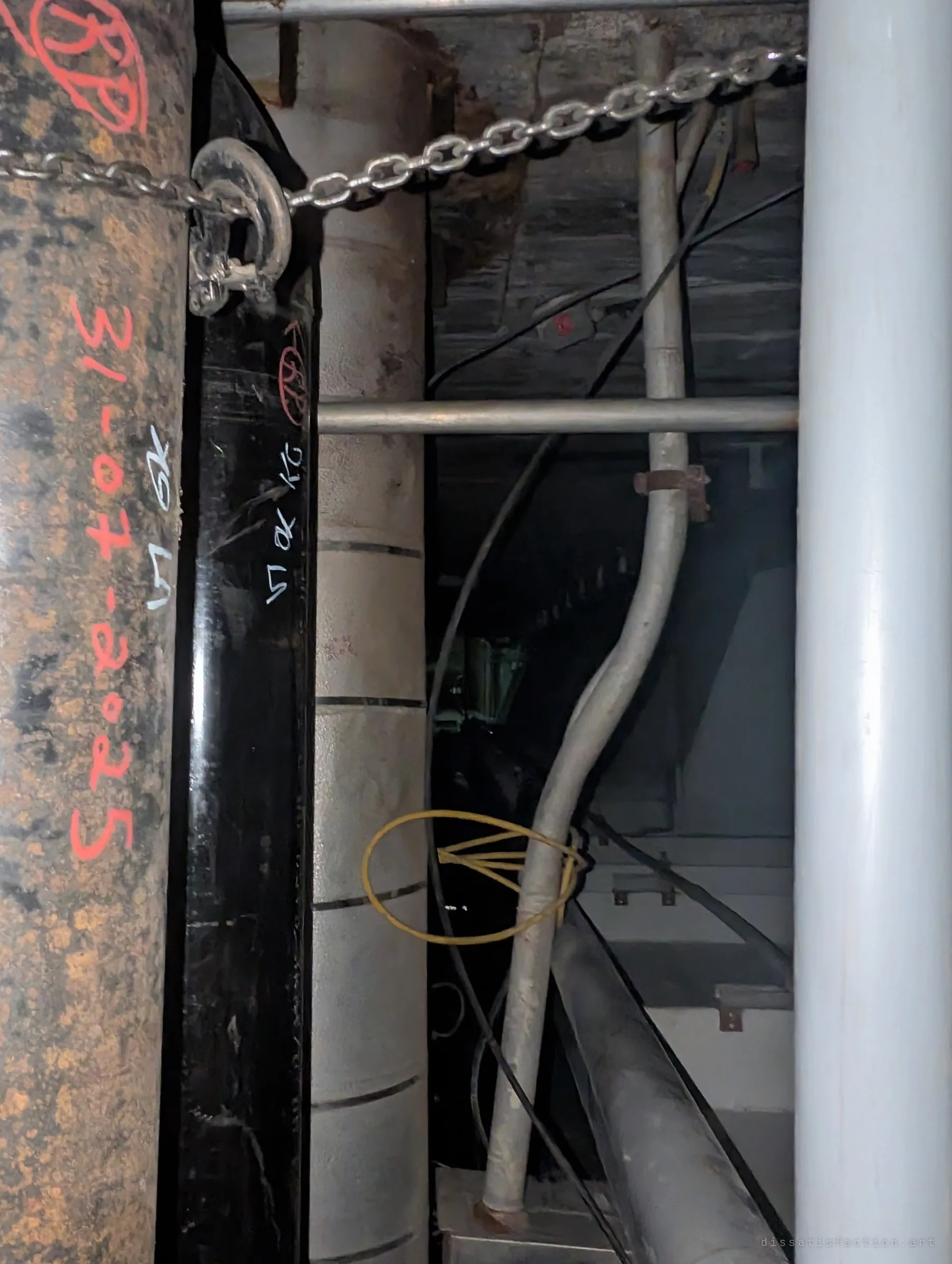 Close-up of new pipes chained in place, marked in red and white paint with inspection stamps and a date, next to insulated risers and conduit. Fresh work threaded through an old structure.