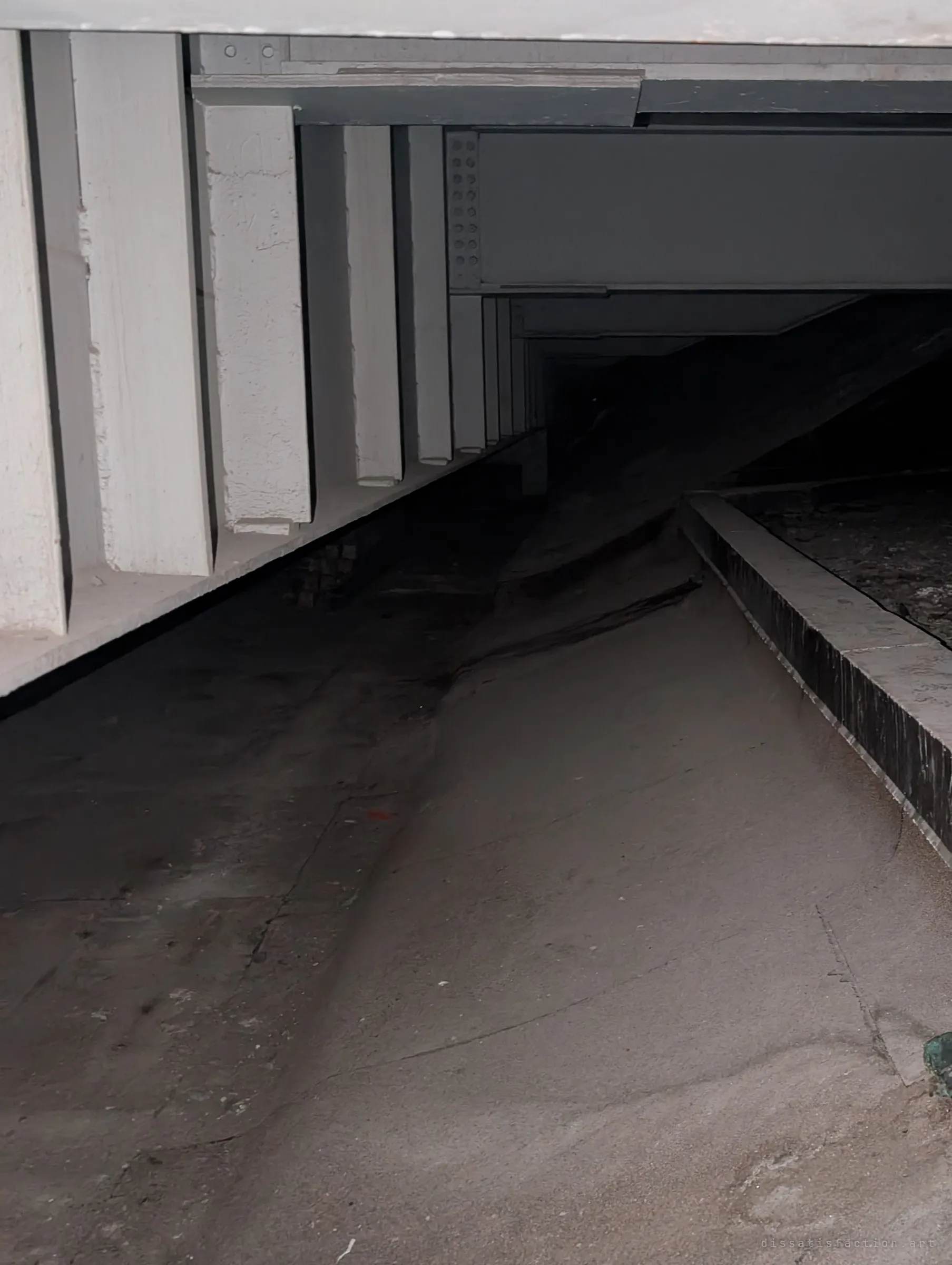 A dusty sloped floor runs under rows of painted concrete and steel beams into darkness. The low ceiling shows how cramped the space beneath the structure is.