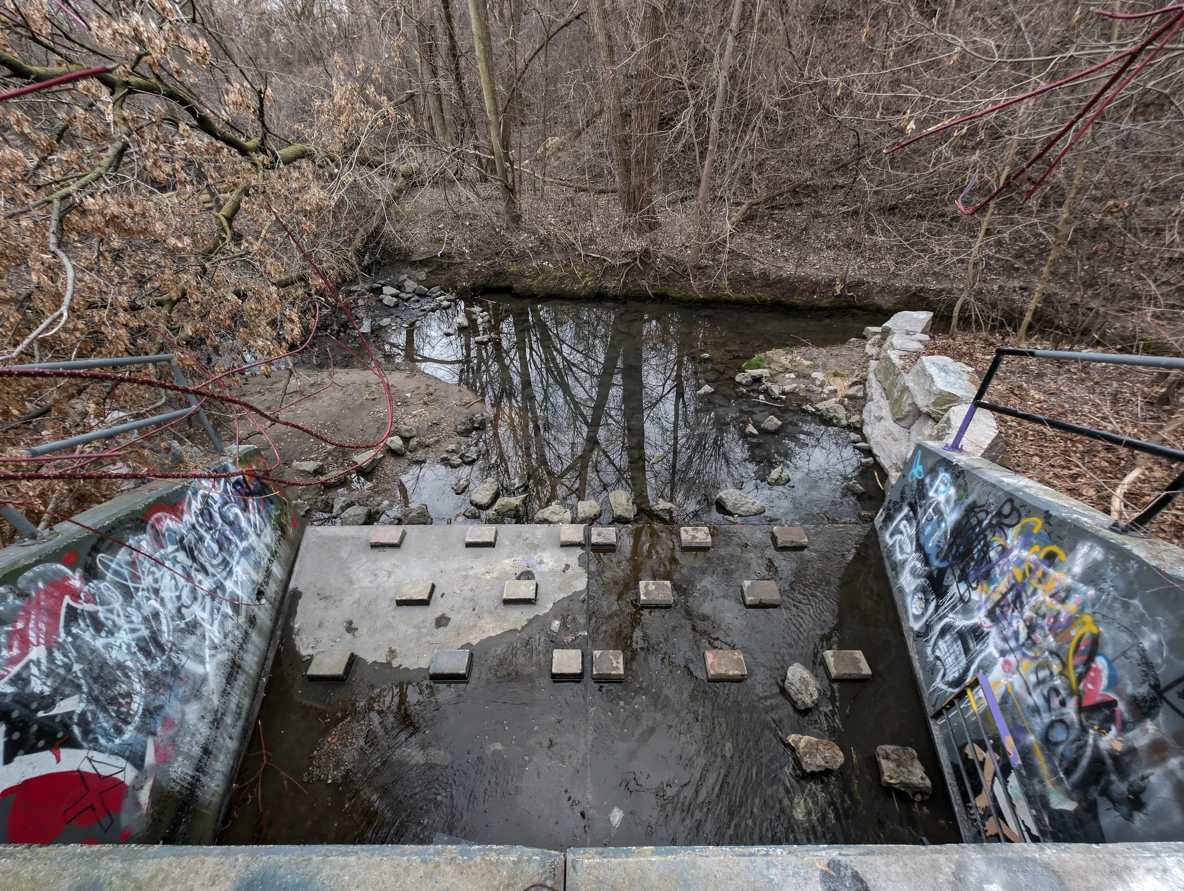 Looking down from the top of a graffiti-covered concrete outfall onto its stepping-block apron and a shallow creek in a bare winter ravine. Sets the scene where the drain empties out.
