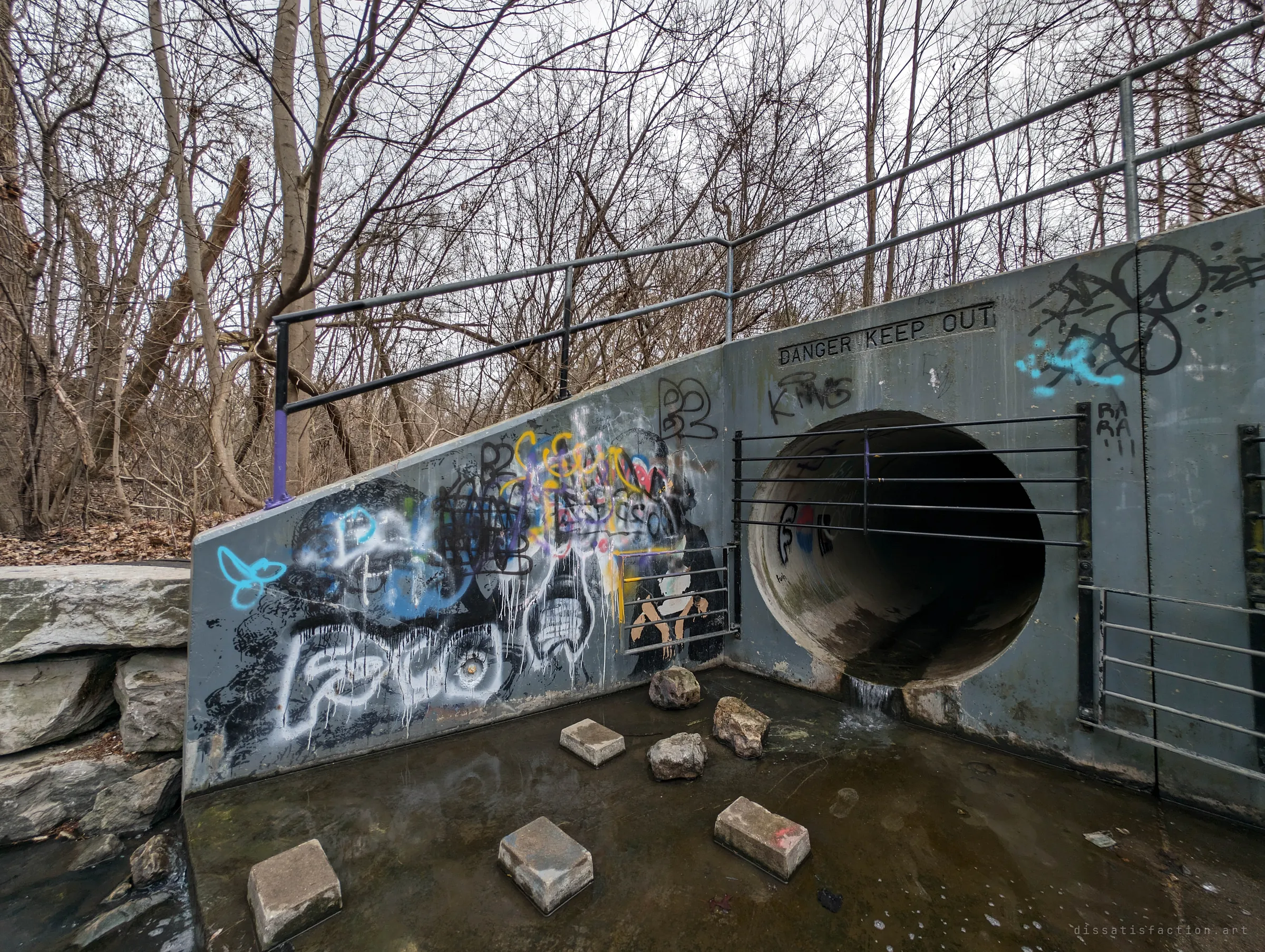 A grated circular drain pipe set into a graffiti-covered concrete headwall with a stock danger warning, stepping blocks in the water below. The way in.