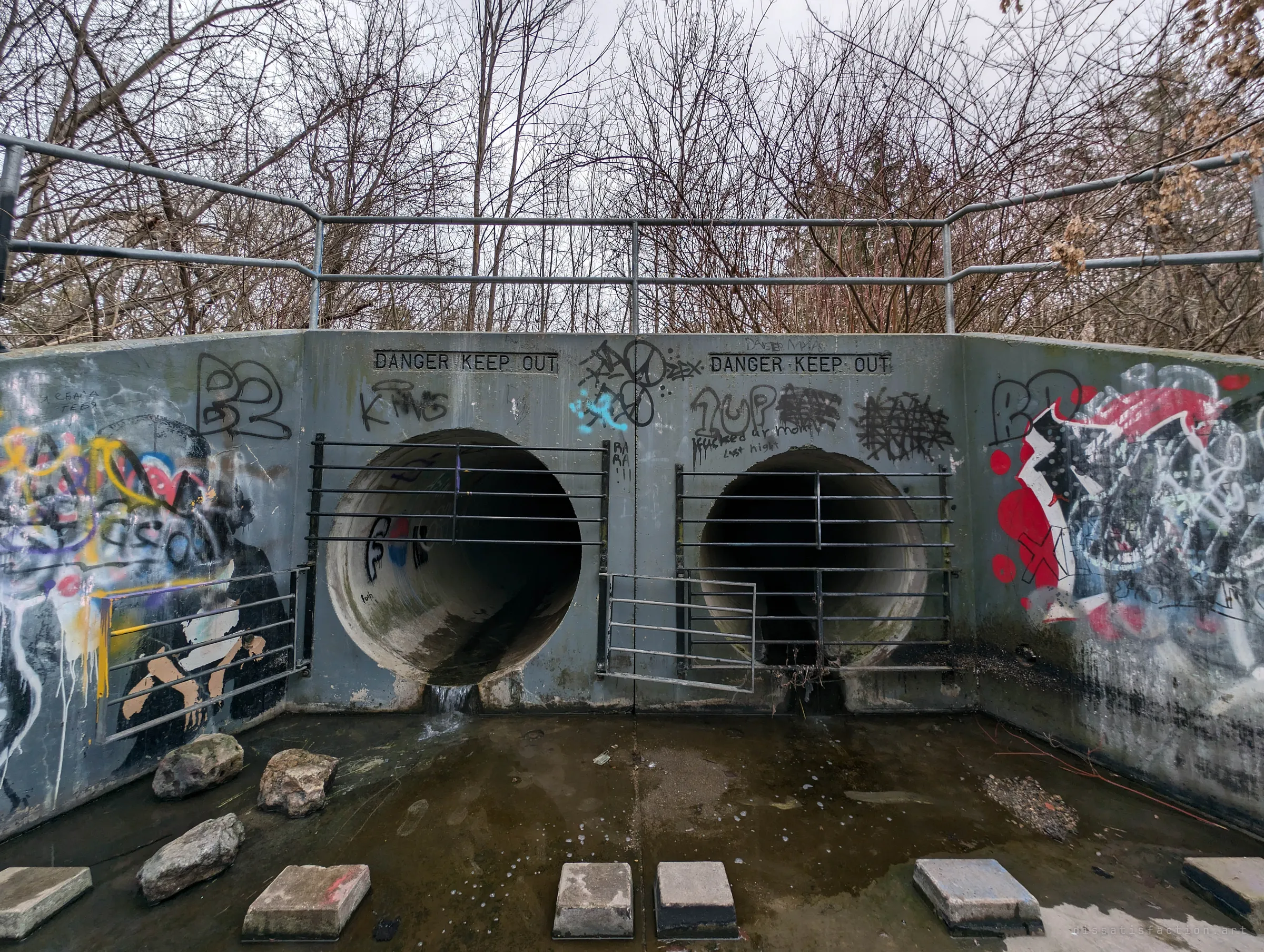 Two barred circular pipes side by side in a graffiti-covered headwall, one gate swung open, water trickling out onto the apron. The front door of the system.
