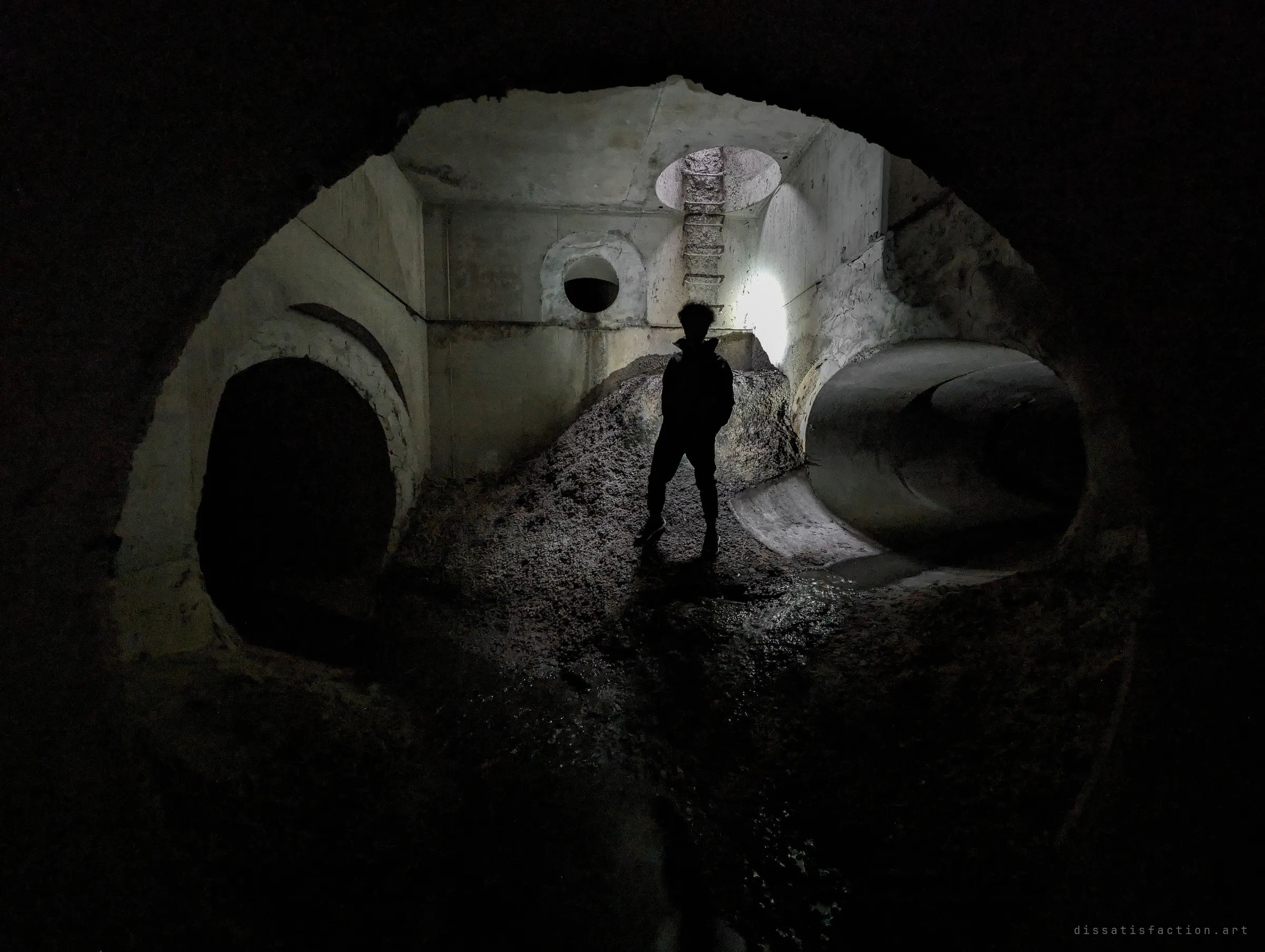 A silhouetted figure stands on a mound of silt in a concrete junction chamber, lit from behind, with pipes branching left and right and a ladder shaft overhead. Shows the scale of the first chamber.