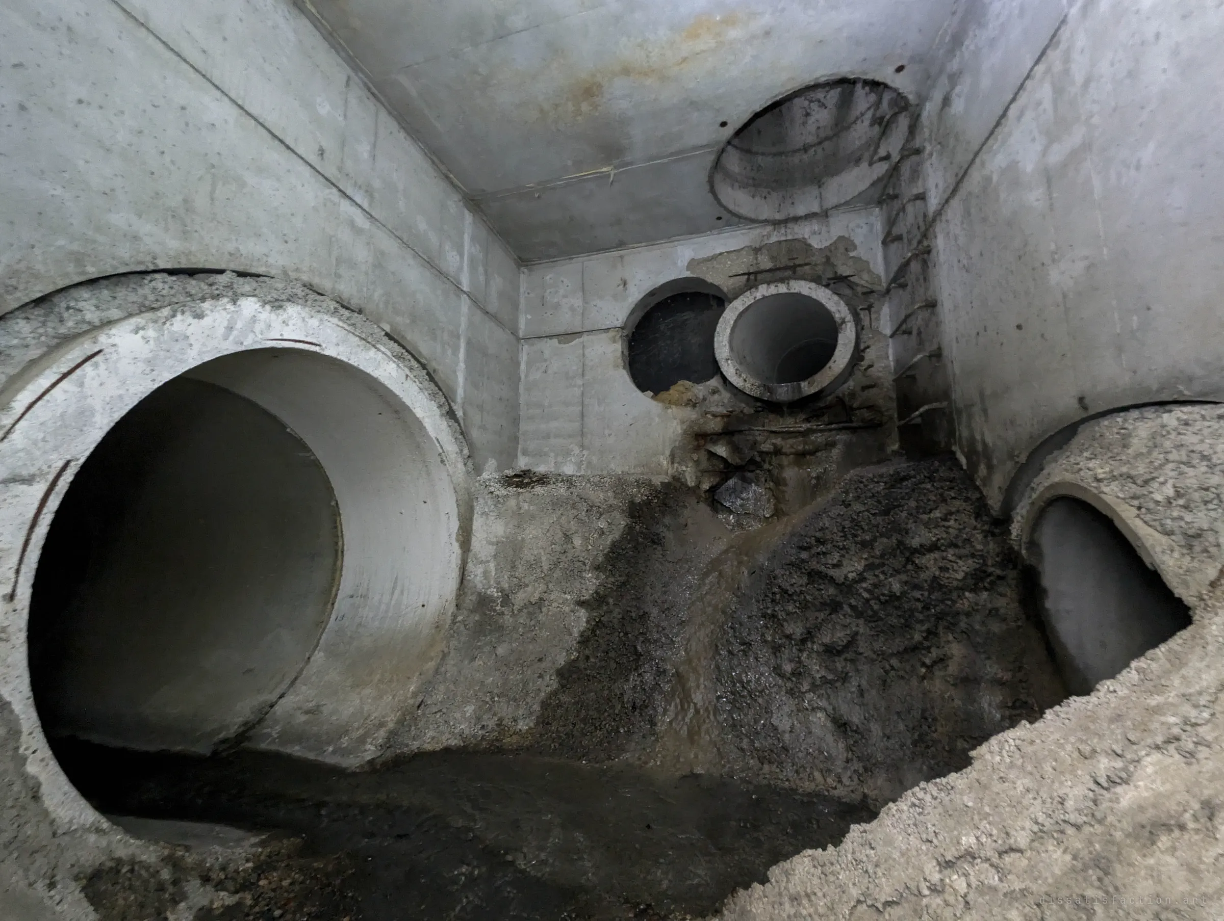 A concrete junction chamber with a large pipe on the left, a smaller broken-in pipe high on the back wall, a manhole shaft above and a slope of silt and debris. Shows how the chamber was pieced together.