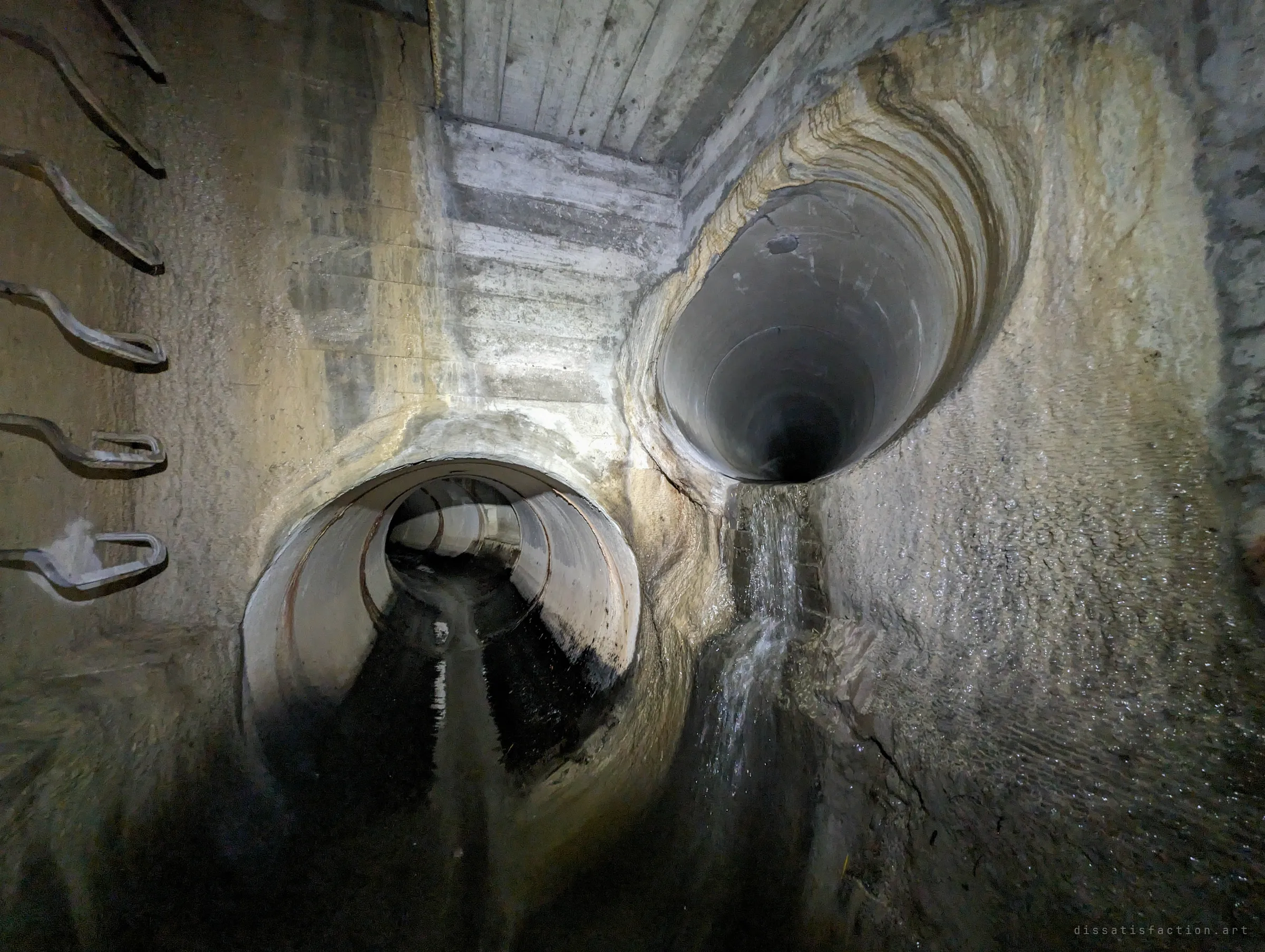 A junction chamber with step irons on the left wall, a round pipe leading away low down and a second pipe high on the wall pouring a thin stream over mineral-stained concrete. It shows how an older branch joins the main line.