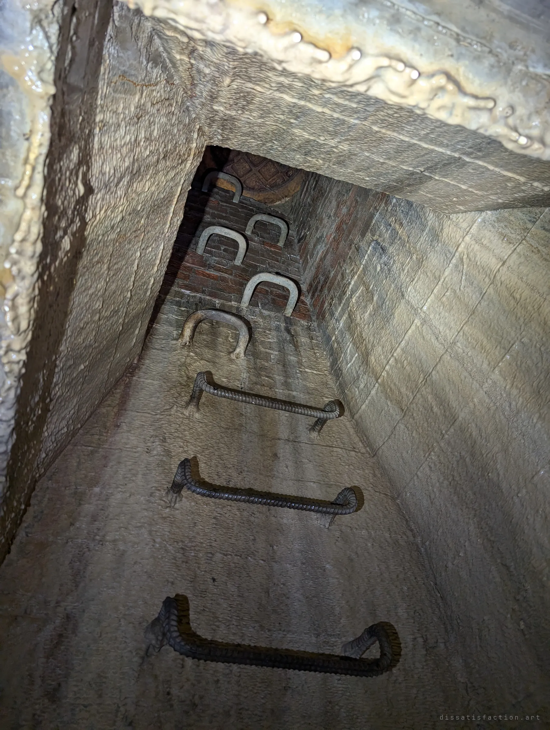 Looking up a narrow access shaft: rebar rungs in concrete give way to older U-shaped step irons set into red brick, ending at a manhole lid. The change in materials shows two eras of construction stacked in one shaft.