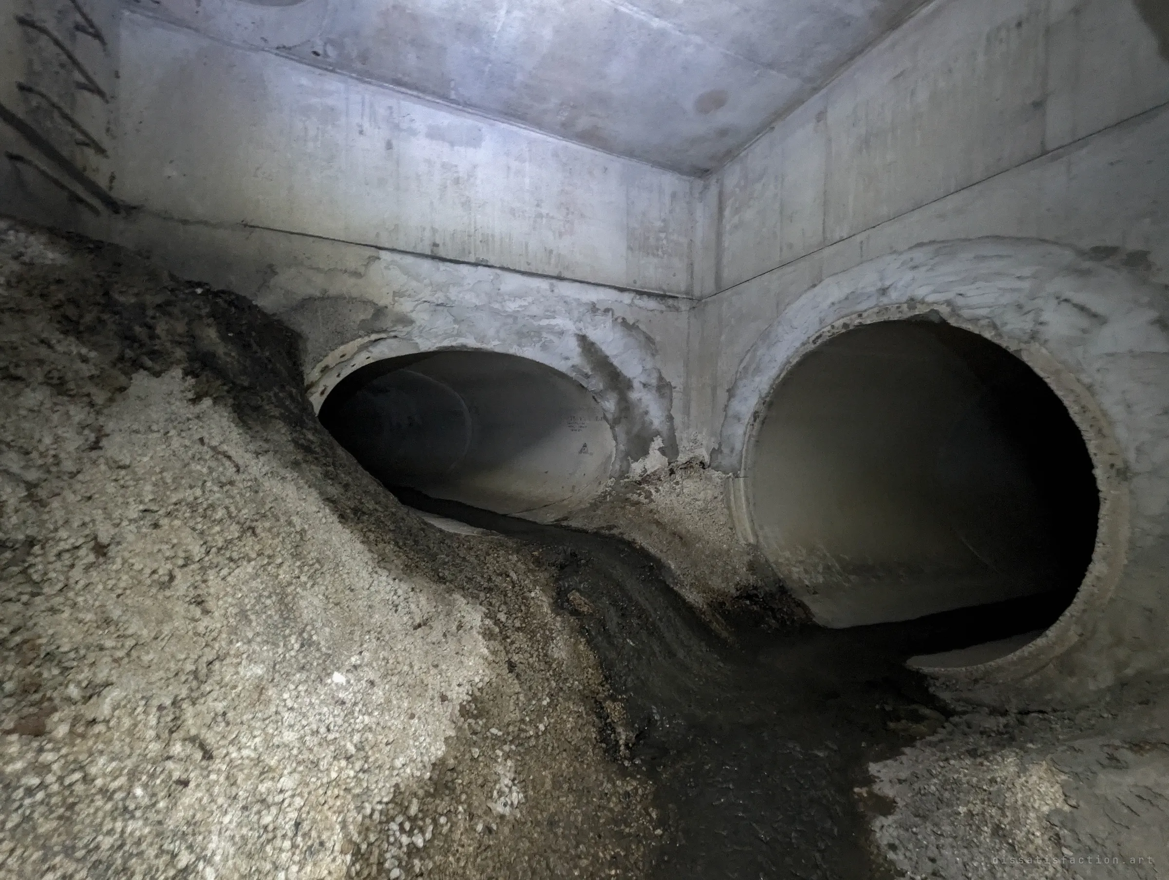 A concrete box chamber where two large round pipes meet, the left one half buried under a bank of gravel with a dark trickle running between them. It shows how much sediment the system has collected.