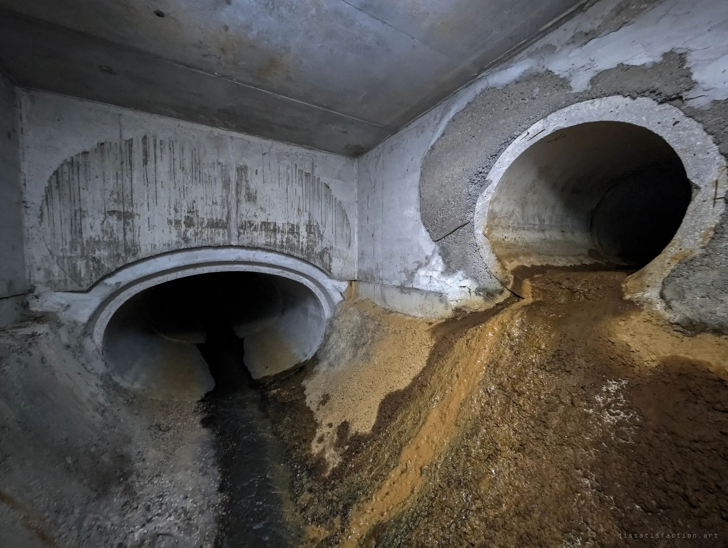 Two pipes enter a concrete chamber side by side, one framed by a flattened arch and the other ringed with rough patching, with orange-stained sediment spilling across the floor. The staining hints at iron-rich water from one branch.
