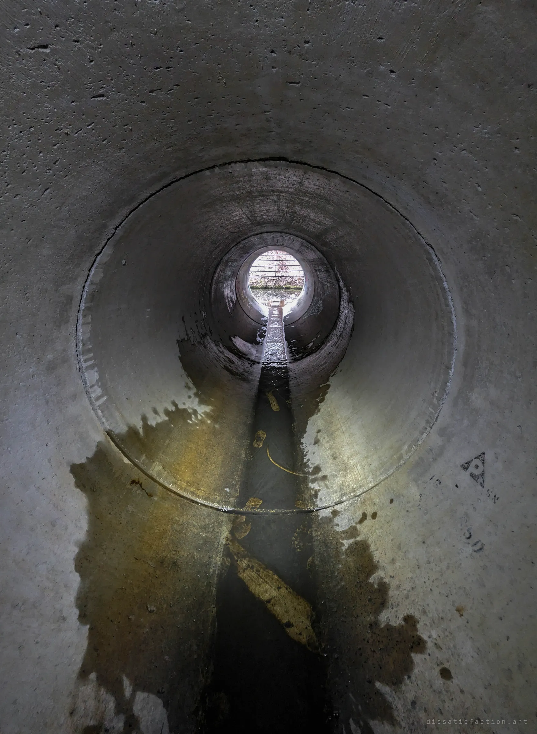 A long round concrete pipe with a narrow low-flow channel running to a bright barred opening at the far end, muddy footprints smeared along the invert. It shows the way out toward daylight.