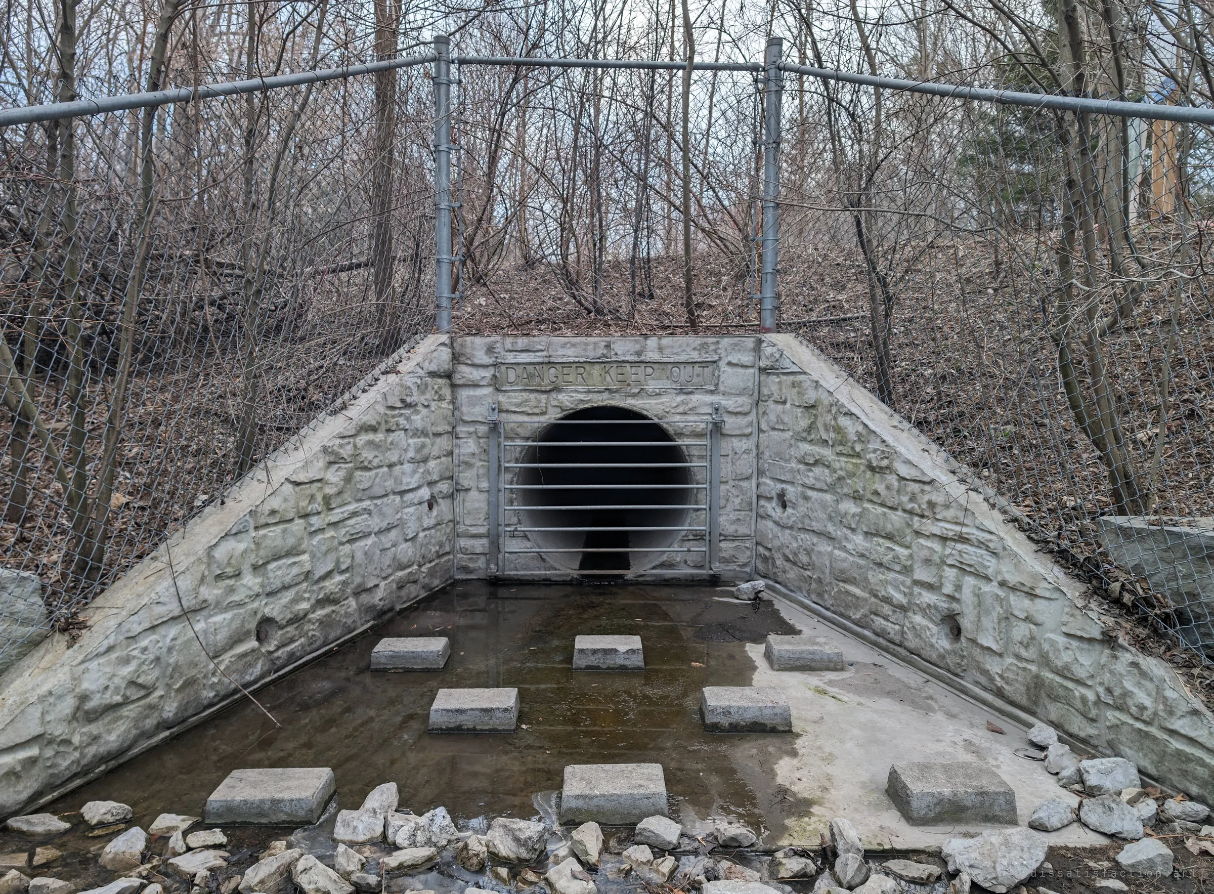 A small barred outfall set in a faux-stone headwall behind a chain-link fence, with concrete blocks standing in shallow water on the apron and bare winter trees above. It shows one of the system's sealed exits from outside.