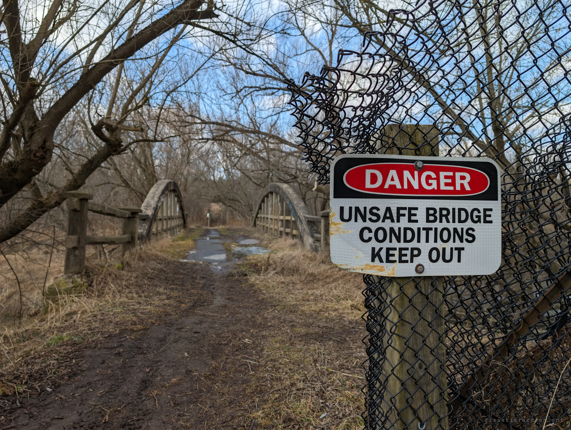 A red and white danger sign on a torn chain-link fence warns of unsafe bridge conditions, with the muddy trail leading onto an old concrete arch bridge behind it. It sets the scene: a crossing officially closed and left to the trees.