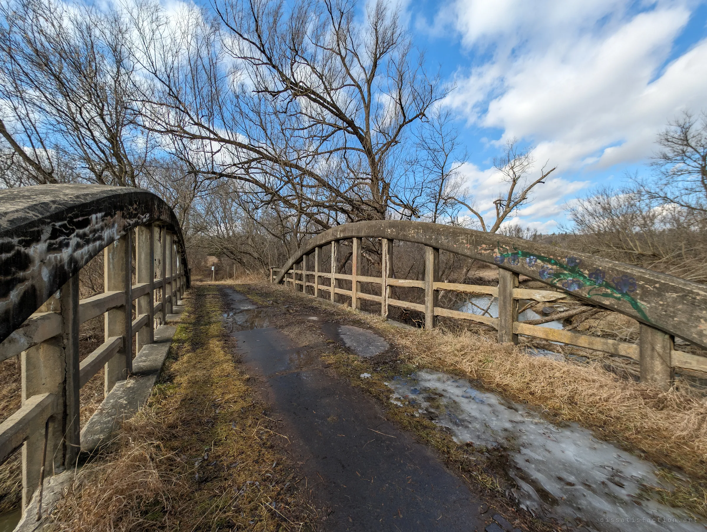 An ultra-wide view across the bridge deck, one arch painted with a vine of blue flowers, the river glinting below under a bright cloud-streaked sky. Ice patches and dead grass cover the old roadway.