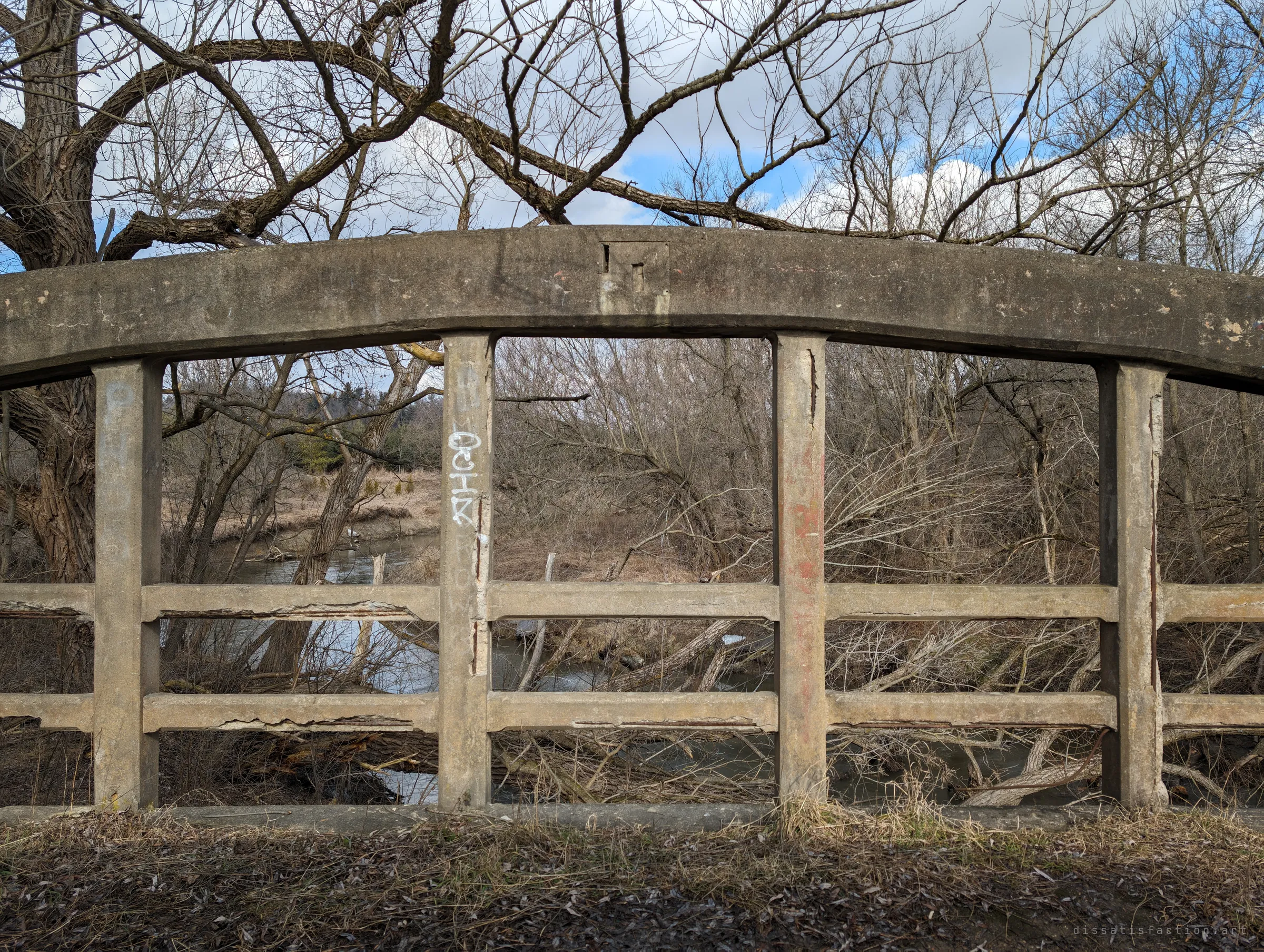 A straight-on view of one arch's concrete railing, its posts cracked and tagged, framing the creek and tangled bare brush beyond. A square notch in the arch hints at a plaque or fitting long since removed.