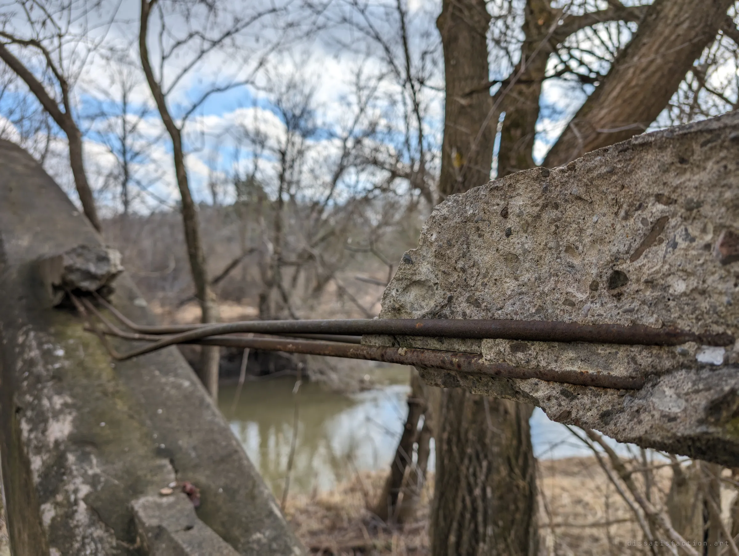 Rusted reinforcing bars bridge a gap where a section of concrete railing has broken away, the river soft and out of focus below. The exposed steel shows how far the structure has decayed.
