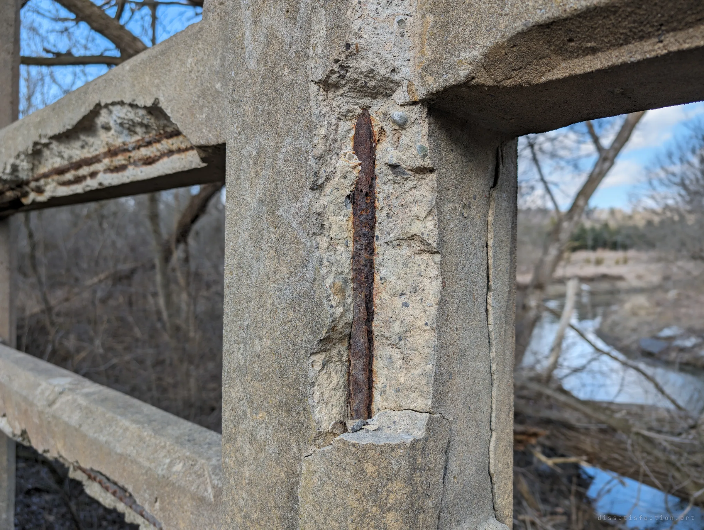 A single corroded rebar is laid bare down the face of a railing post, with more broken concrete on the rails beside it and the creek behind. Detail of the slow failure holding the bridge together.