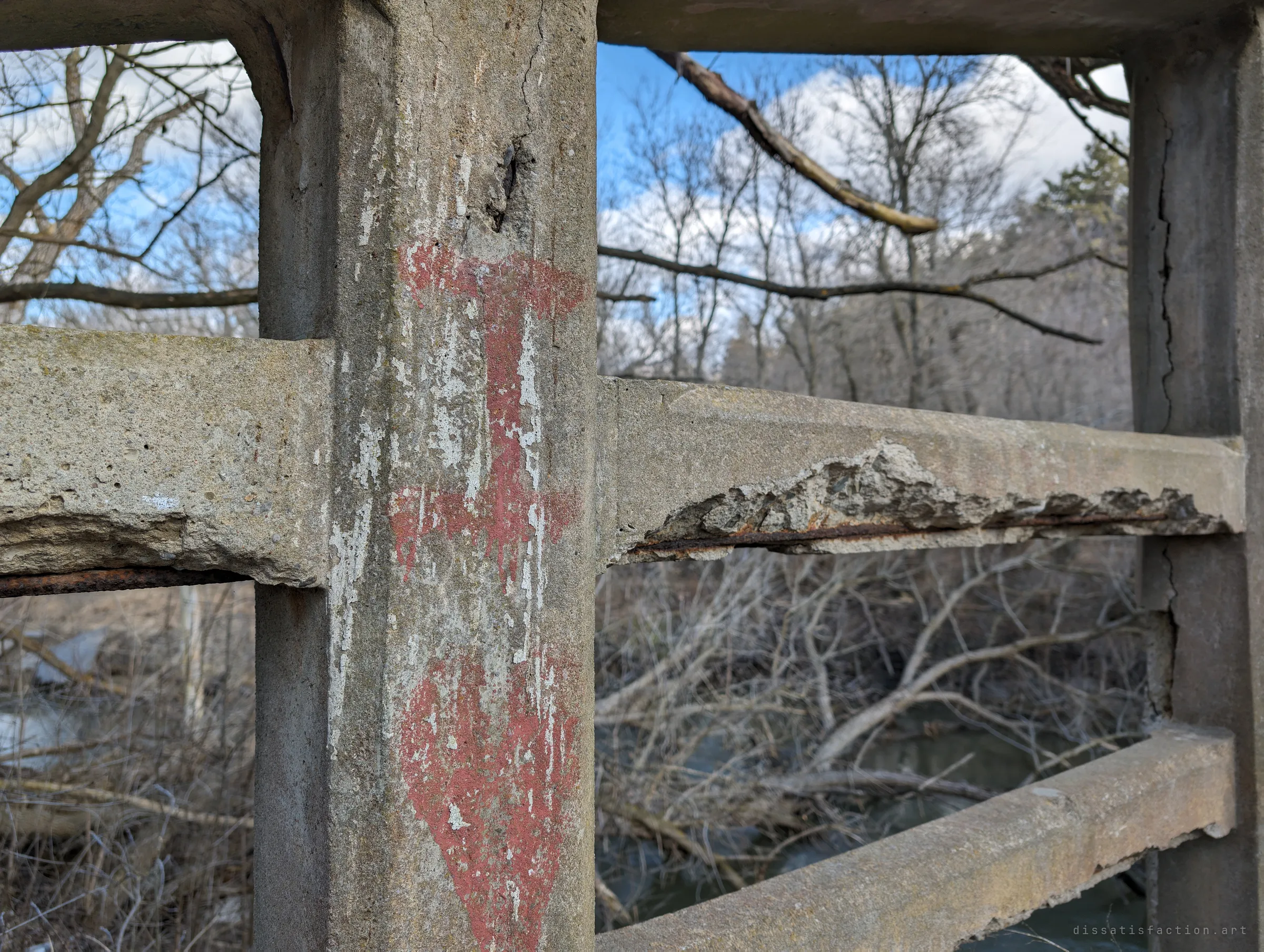 Faded red paint drips down a cracked railing post while the rail beside it has crumbled to expose its steel. The creek runs through fallen branches below.