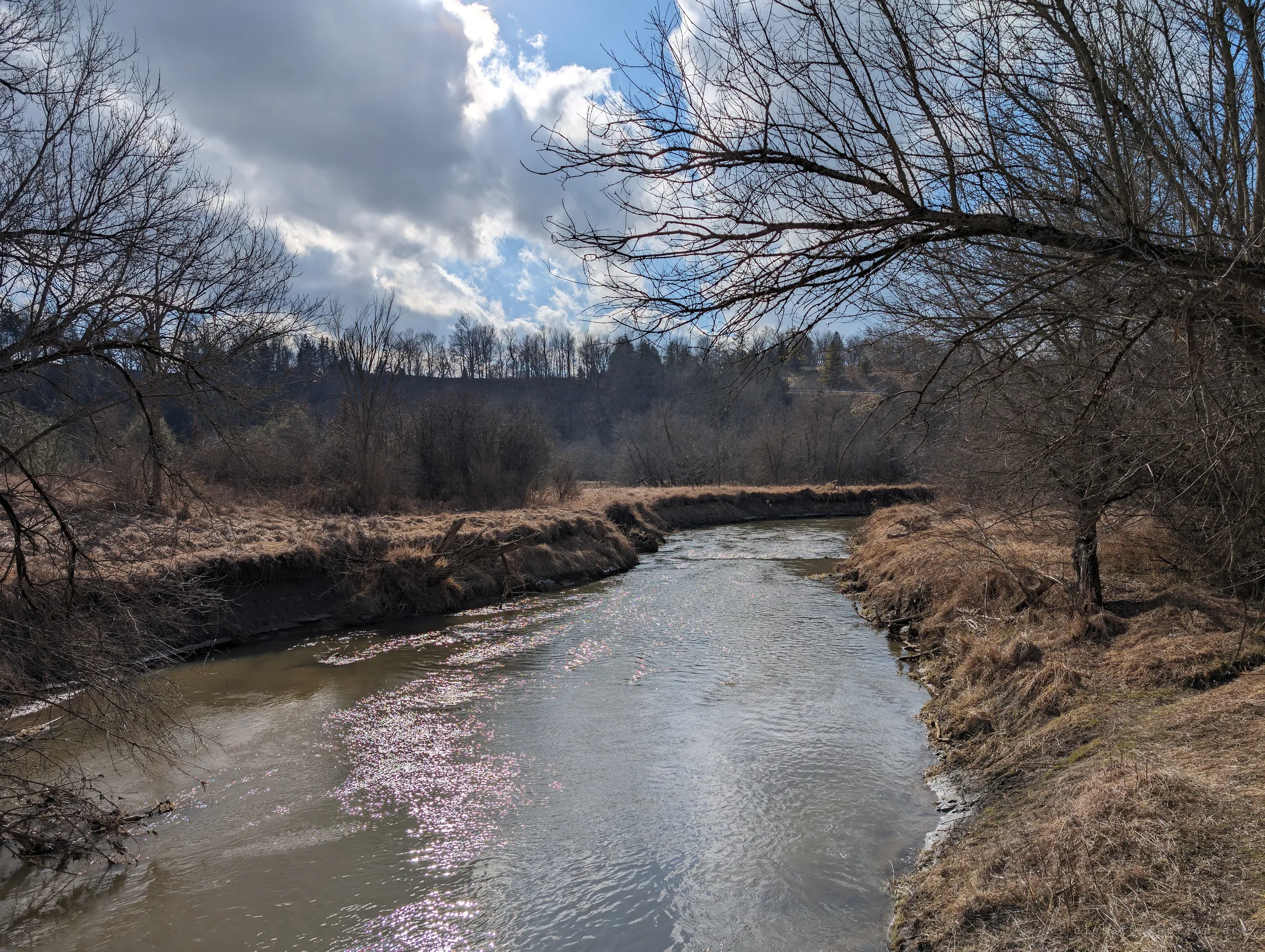 The creek bends away between grassy banks under a moody sky, sunlight sparkling on the water. The quiet valley the old bridge once carried traffic across.