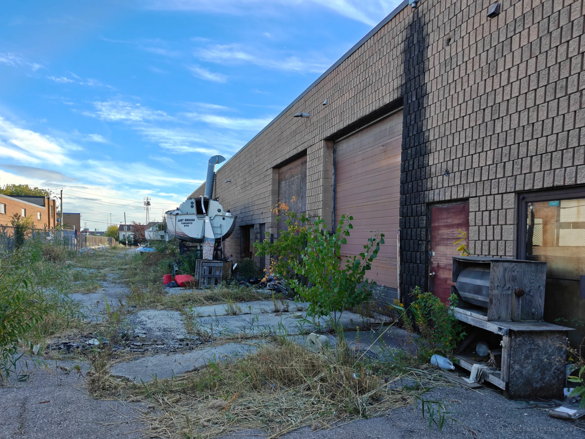 The back lot of a block-walled unit, weeds pushing through broken asphalt toward a sailboat left on its stand beside shuttered loading doors. Smoke stains streak the wall above one of the doors.