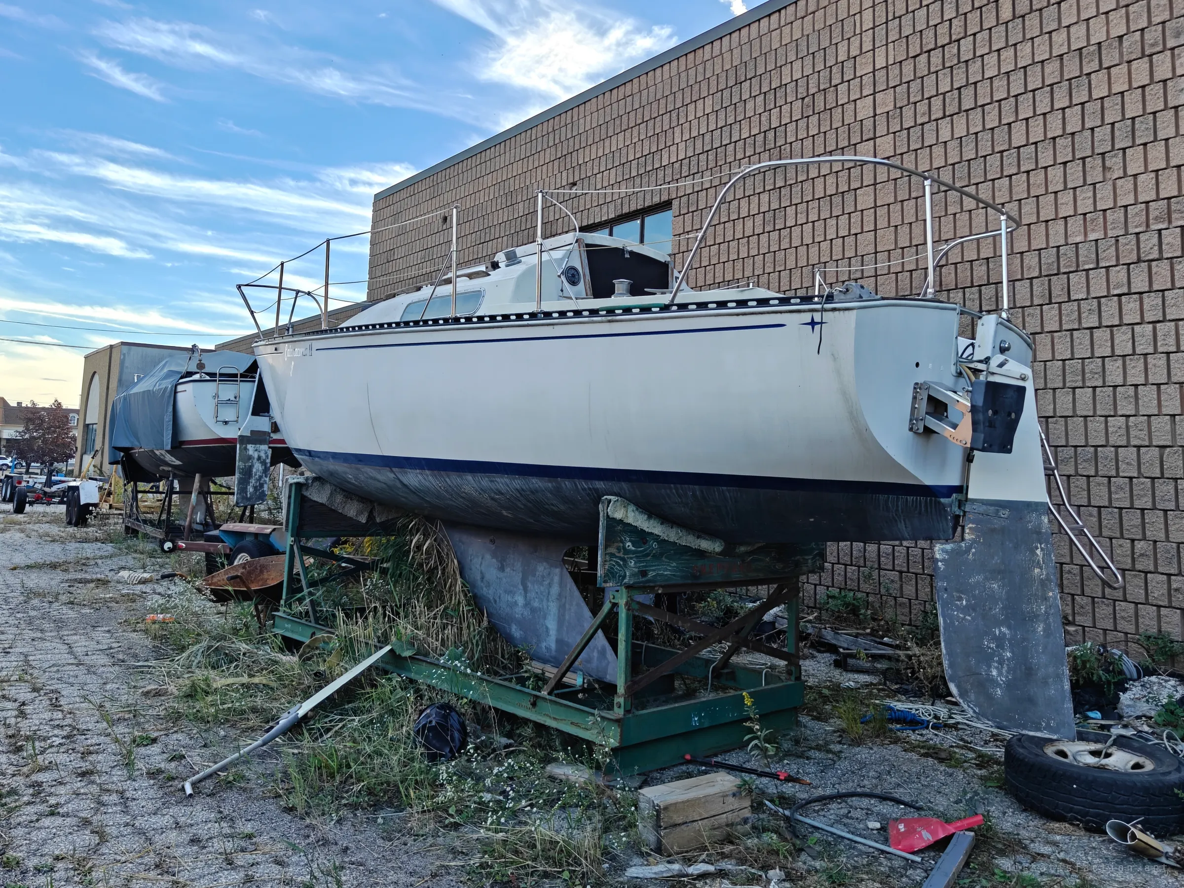 A small sailboat sits propped in a green steel cradle, grass growing up through the frame and its rudder hanging loose. More boats and trailers line the wall behind it.