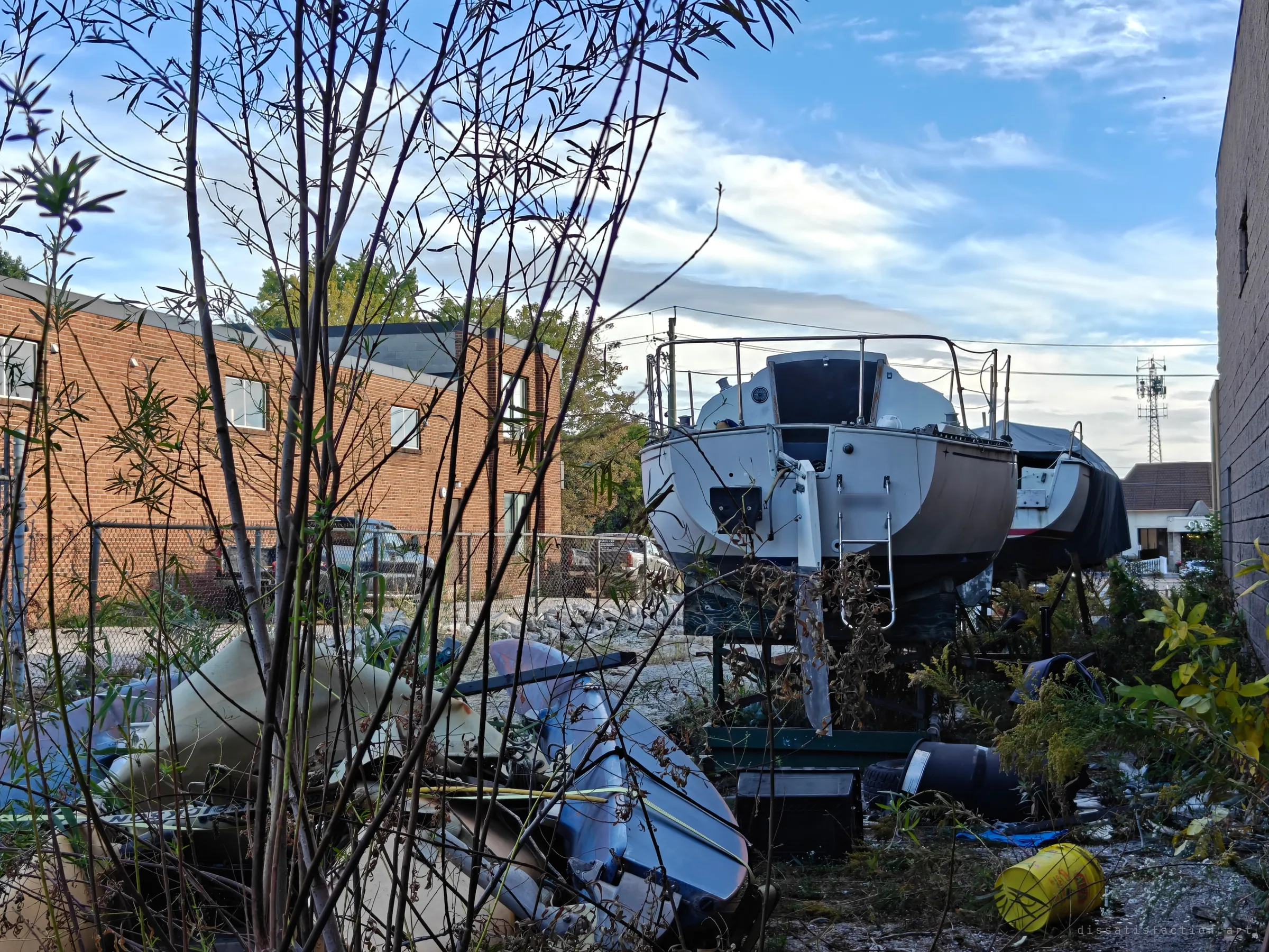 Seen through bare saplings, a sailboat's stern faces a heap of broken fibreglass and debris. The yard has become a slow graveyard for boats nobody came back for.
