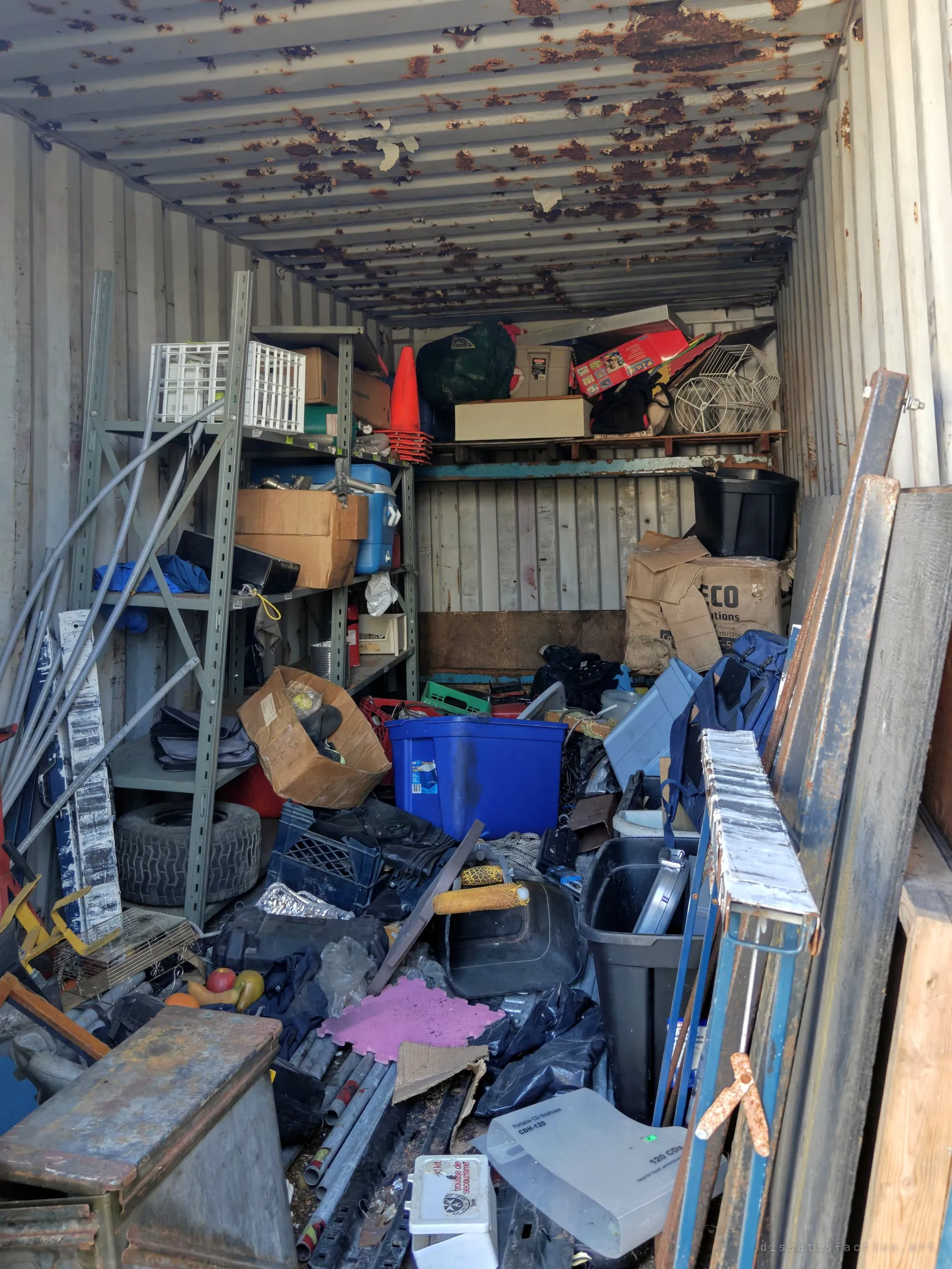 Inside a shipping container with a rust-blistered ceiling, shelves and bins are buried under tools, crates, pipes and boxes. It looks like years of storage that nobody ever sorted.