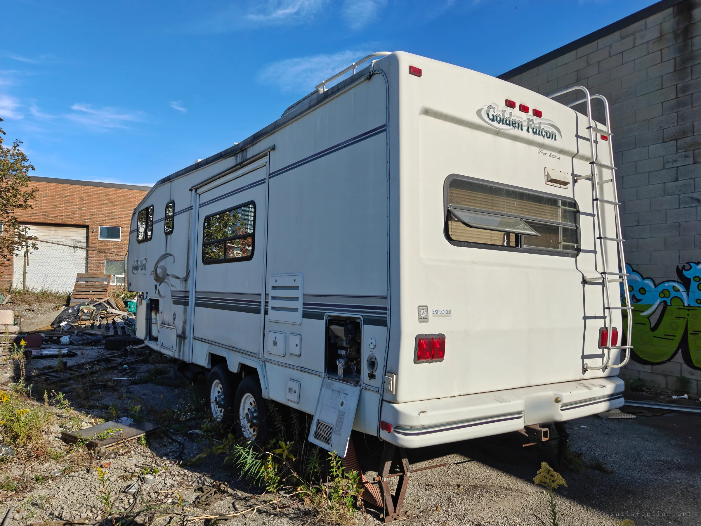 A fifth-wheel camper sits abandoned in the lot, its utility hatch hanging open and weeds growing around the wheels. Pallets and scrap are piled beside it.