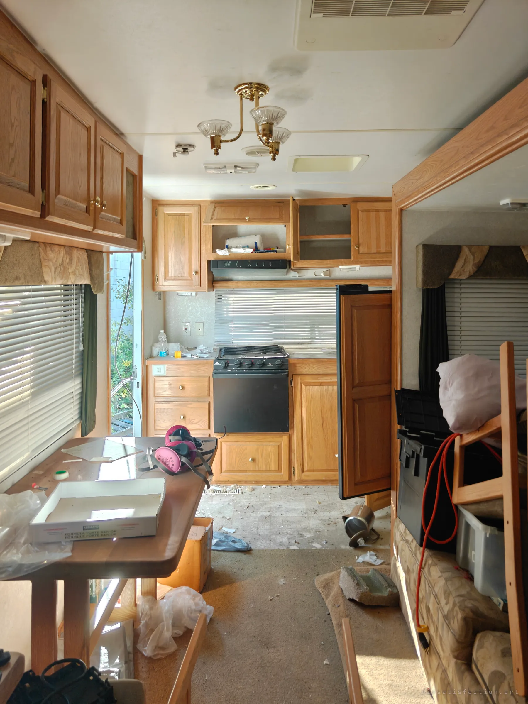 Looking down the length of an abandoned travel trailer: oak cabinets, a brass ceiling fixture, respirator masks on the dinette table and debris across the floor. The open door lets daylight spill into the stripped-out interior.
