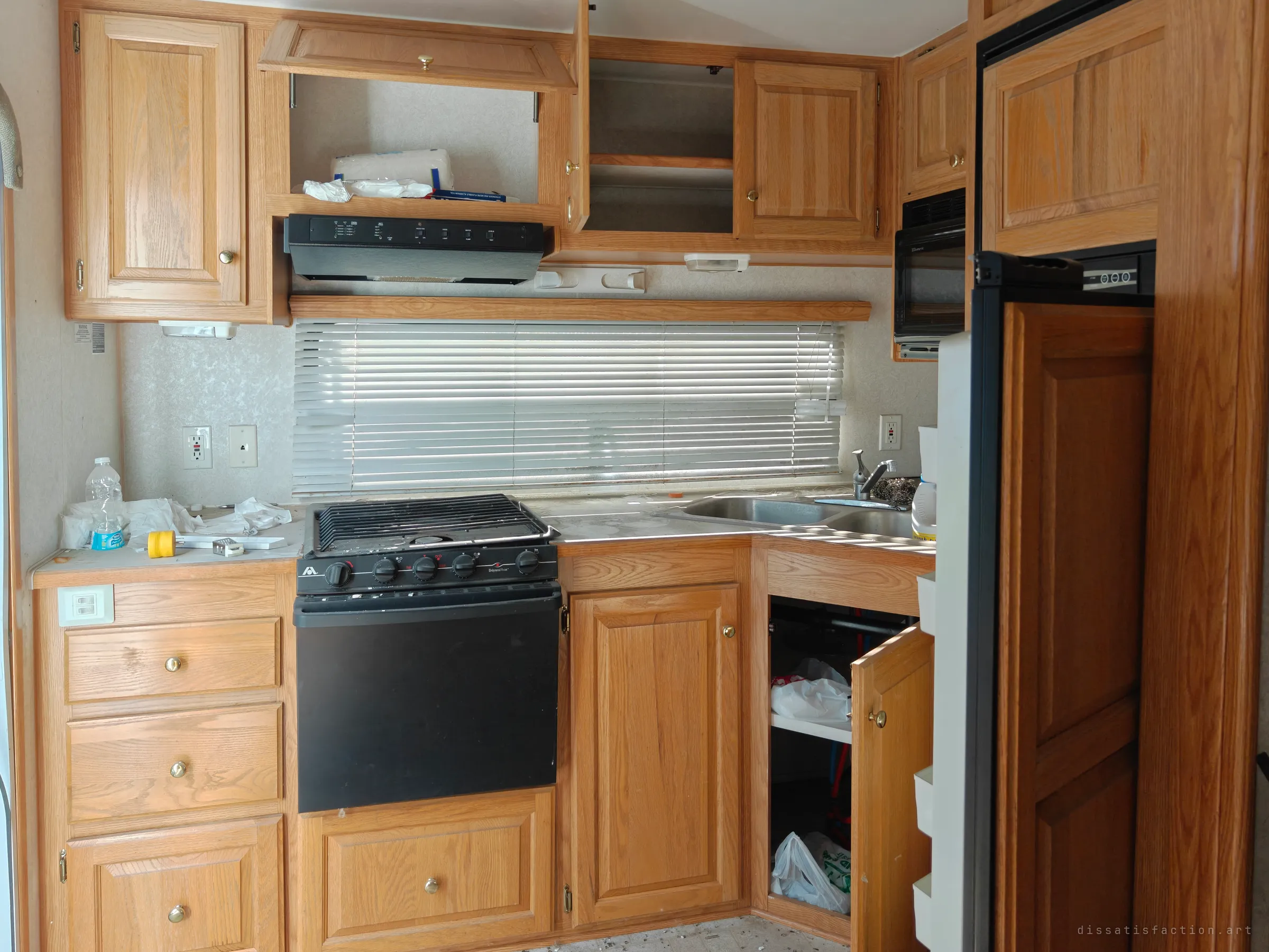 The trailer's galley kitchen, with oak cabinets hanging open, a black stove and a dusty counter under closed blinds. Everything looks left mid-use, from the water bottle to the scattered paper towels.