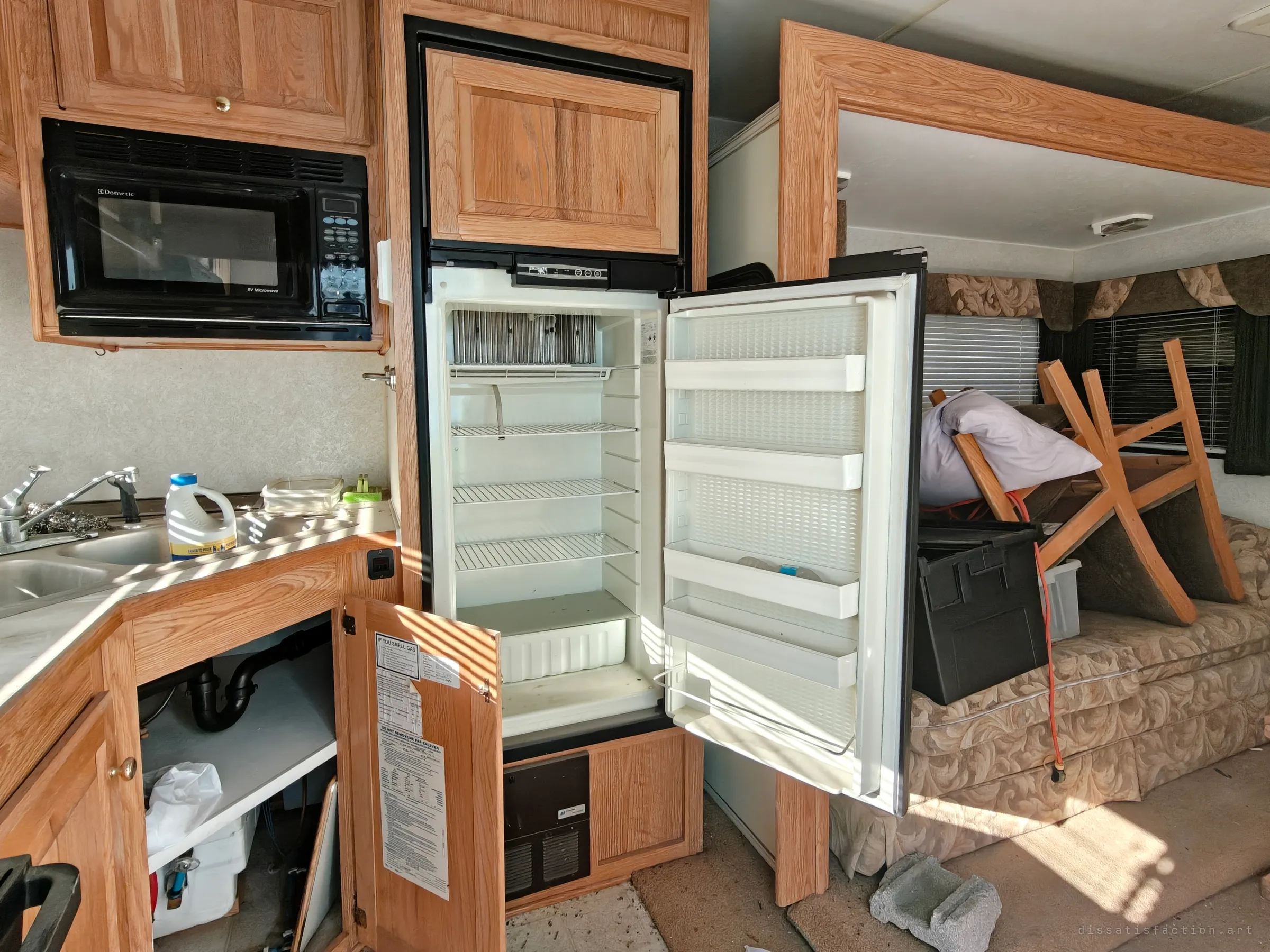 An empty fridge stands open beside a built-in microwave, with sunlight striping the counter through the blinds. In the living area behind, a chair is piled upside down on the sofa with bins and an extension cord.