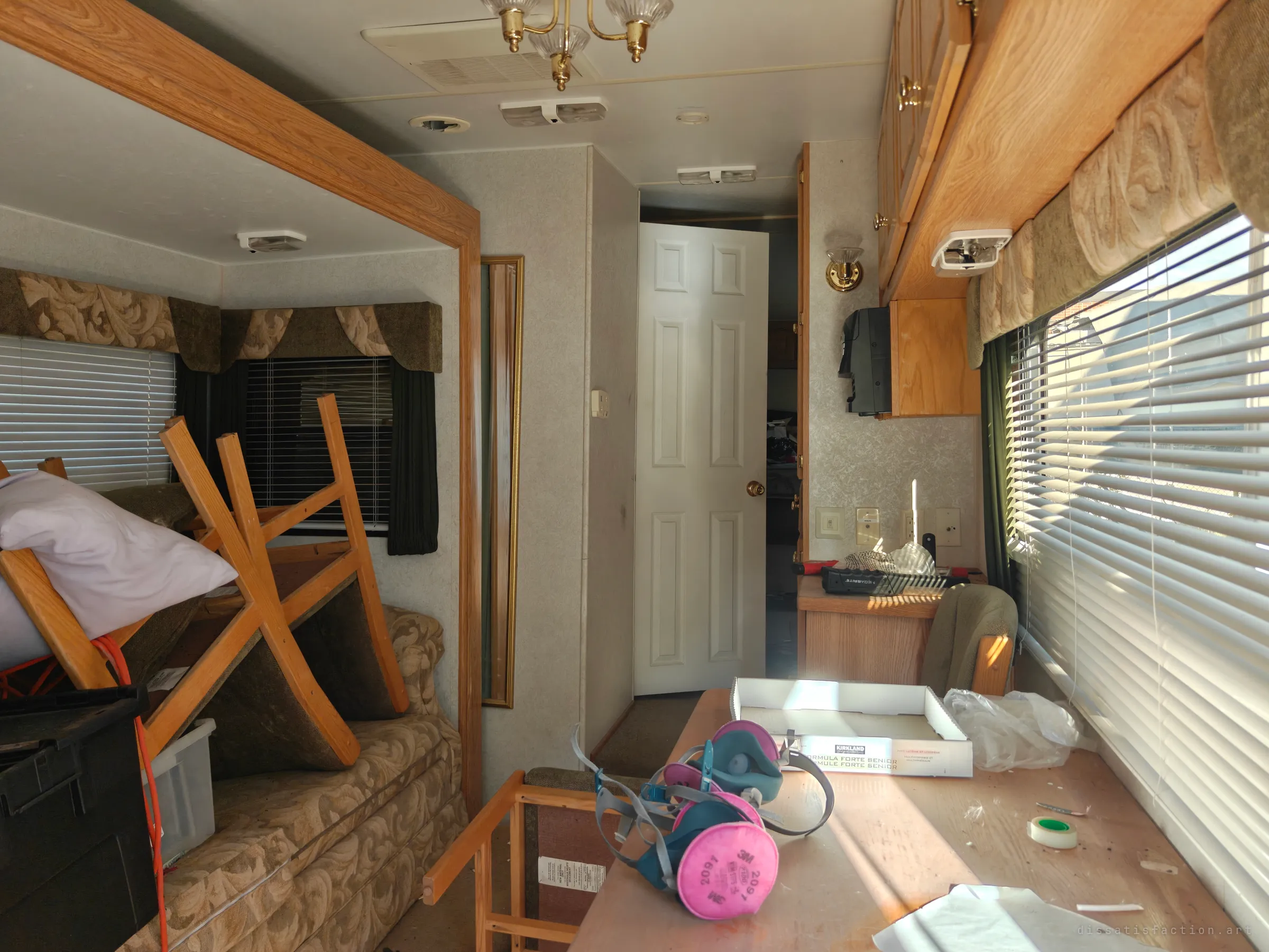 The trailer's living area seen from the dinette, with respirator masks and an empty box on the table and furniture stacked on the sofa. A white interior door stands open toward the bedroom at the back.