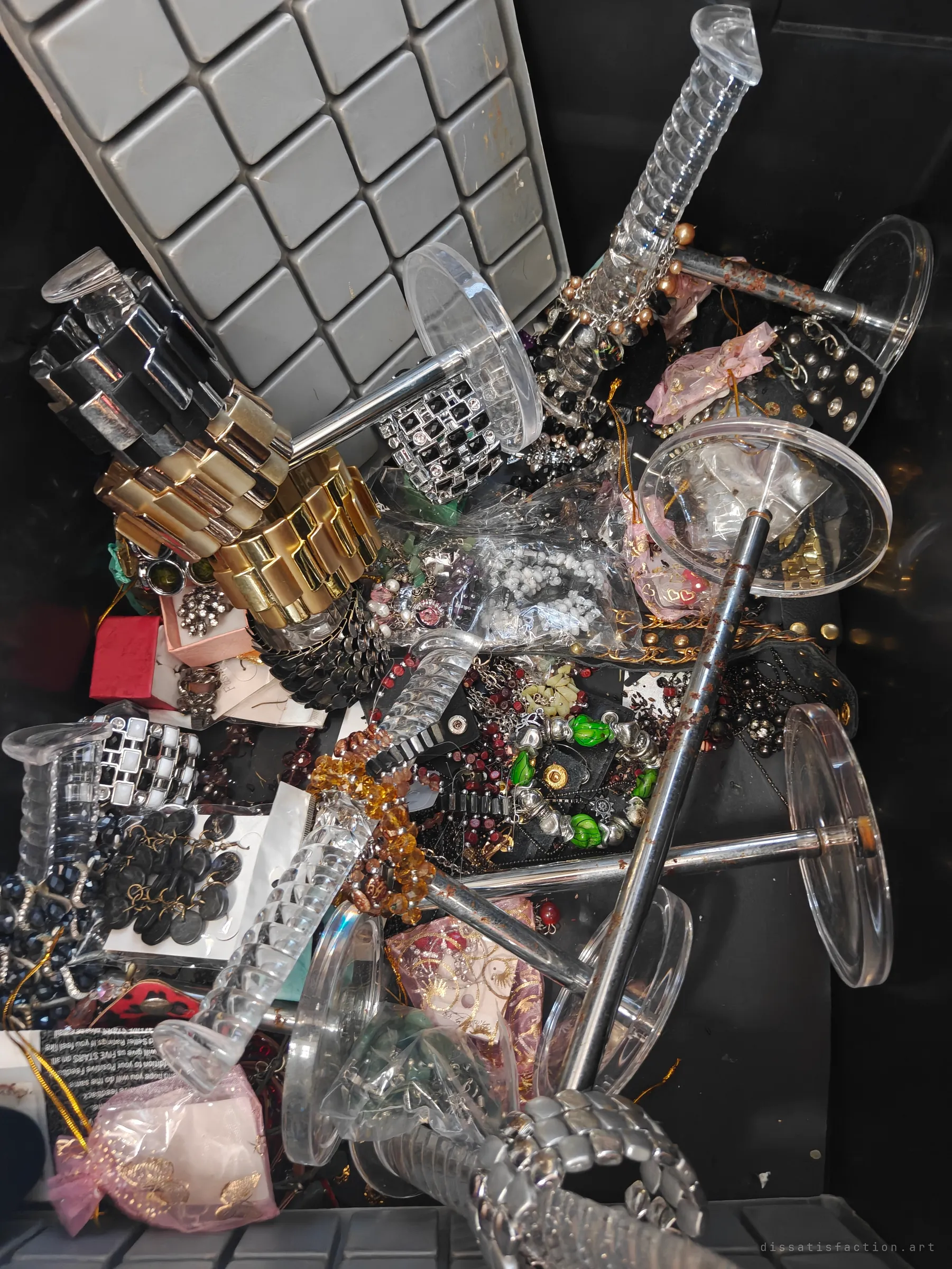A bin crammed with costume jewellery: chunky gold and silver bracelets, beaded strands, earrings on cards and clear acrylic display stands tangled together. It looks like a whole shop's stock was dumped at once.
